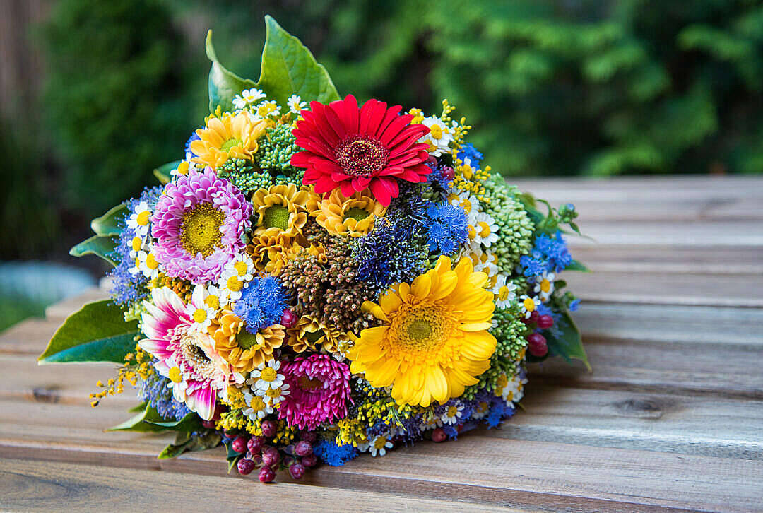 花朵 4K 桌布 壁紙 — 一束鮮豔的紅、粉紅、黃和藍色花朵放在一張飽經風霜的木桌上，背景是柔和的綠色。