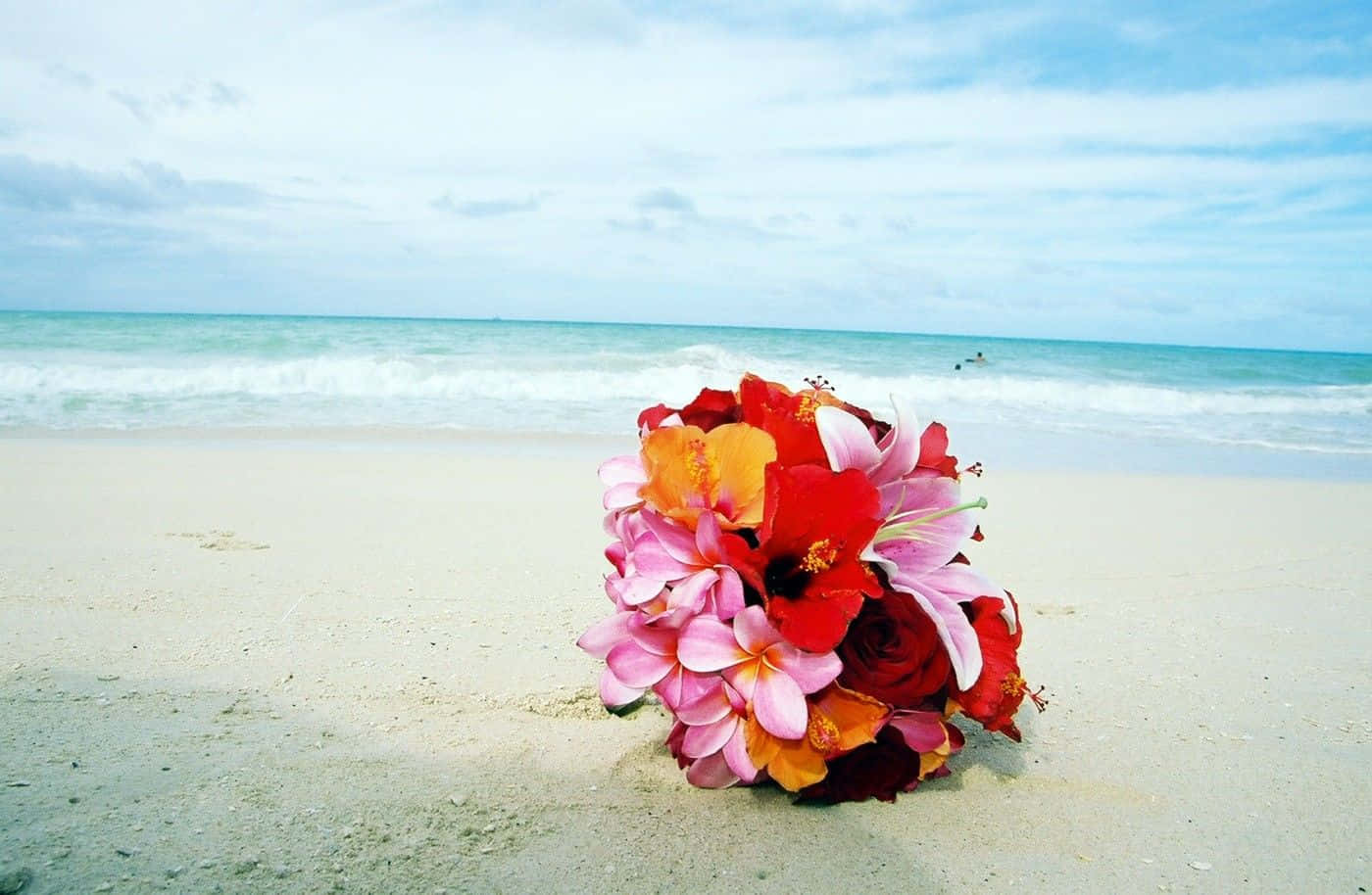 Hawaiian Beach 桌布 壁紙 — 一束鮮豔的紅、粉紅和橙色花朵放在沙灘上，背景有海浪和陰天。