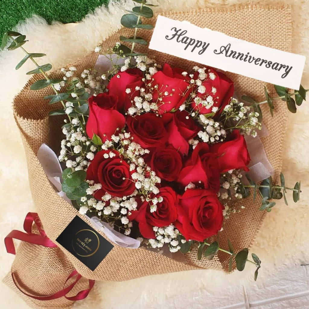 Rose Flower 桌布 壁紙 — 一束鮮豔的紅玫瑰和滿天星，用麻布包裹，繫上「Happy Anniversary」標籤，放在柔軟的白色毛絨表面上。