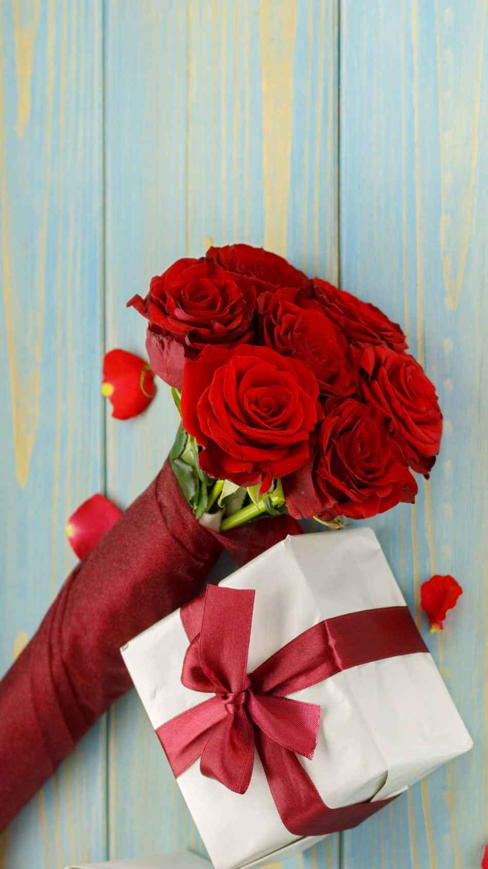 Valentines 桌布 壁紙 — 一束鮮豔的紅玫瑰放在酒紅色袖套中，旁邊是一個用相配紅色絲帶綁著的白色禮物盒，放在風化的藍色木質表面上。
