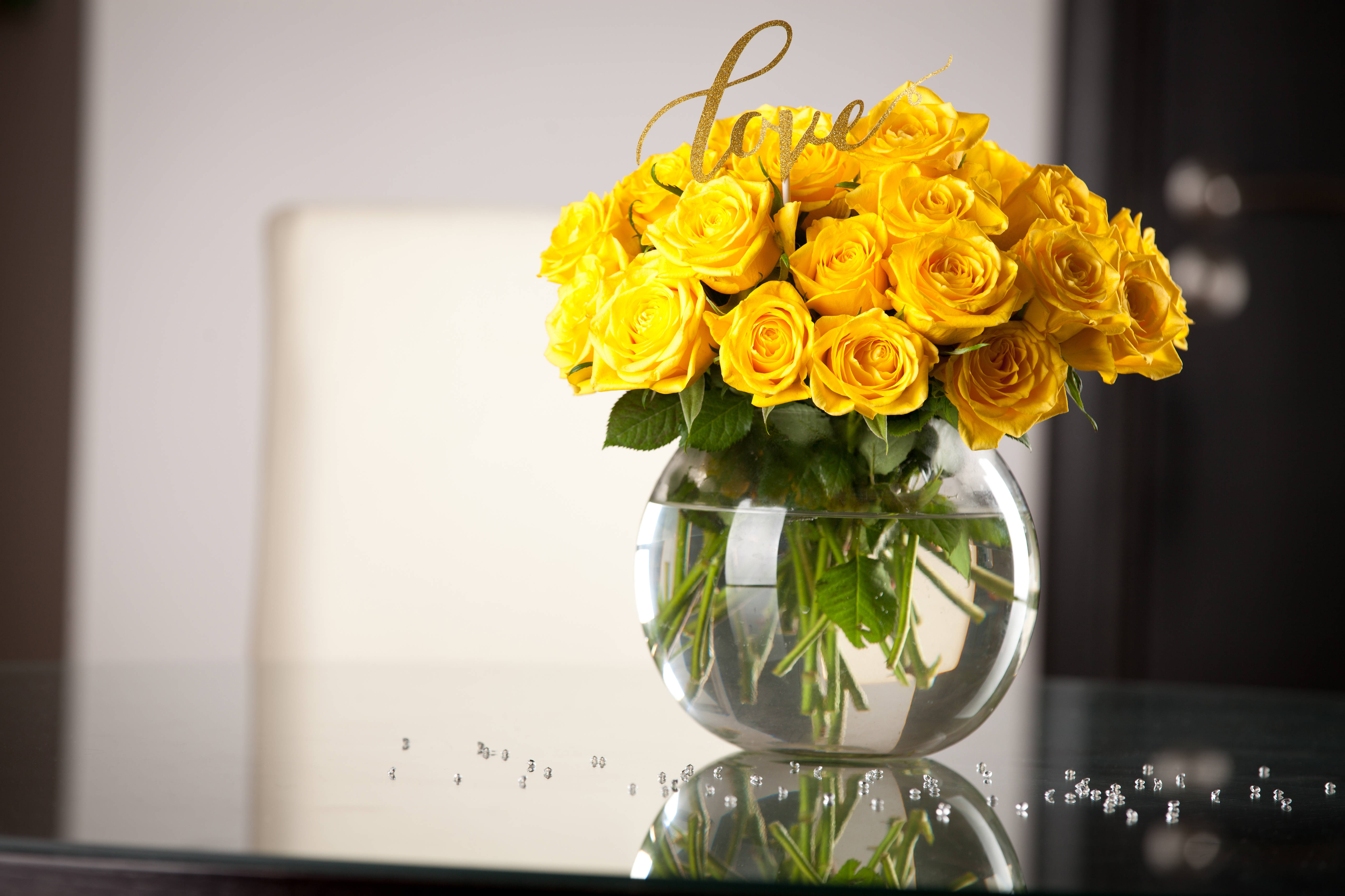 Flowers 桌布 壁紙 — 一束鮮豔的黃玫瑰插在透明玻璃花瓶中，放在反光桌上，帶有金色的 'love' 裝飾。