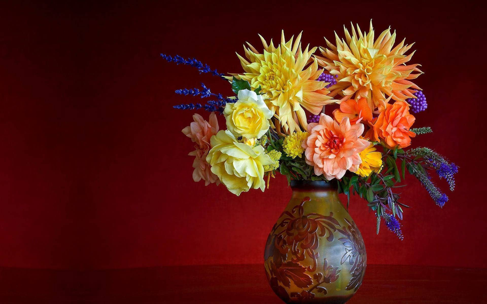 Flowers 桌布 壁紙 — 一束鮮豔的黃色、橙色和粉紅色花朵，插在帶圖案的花瓶中，背景為深紅色。
