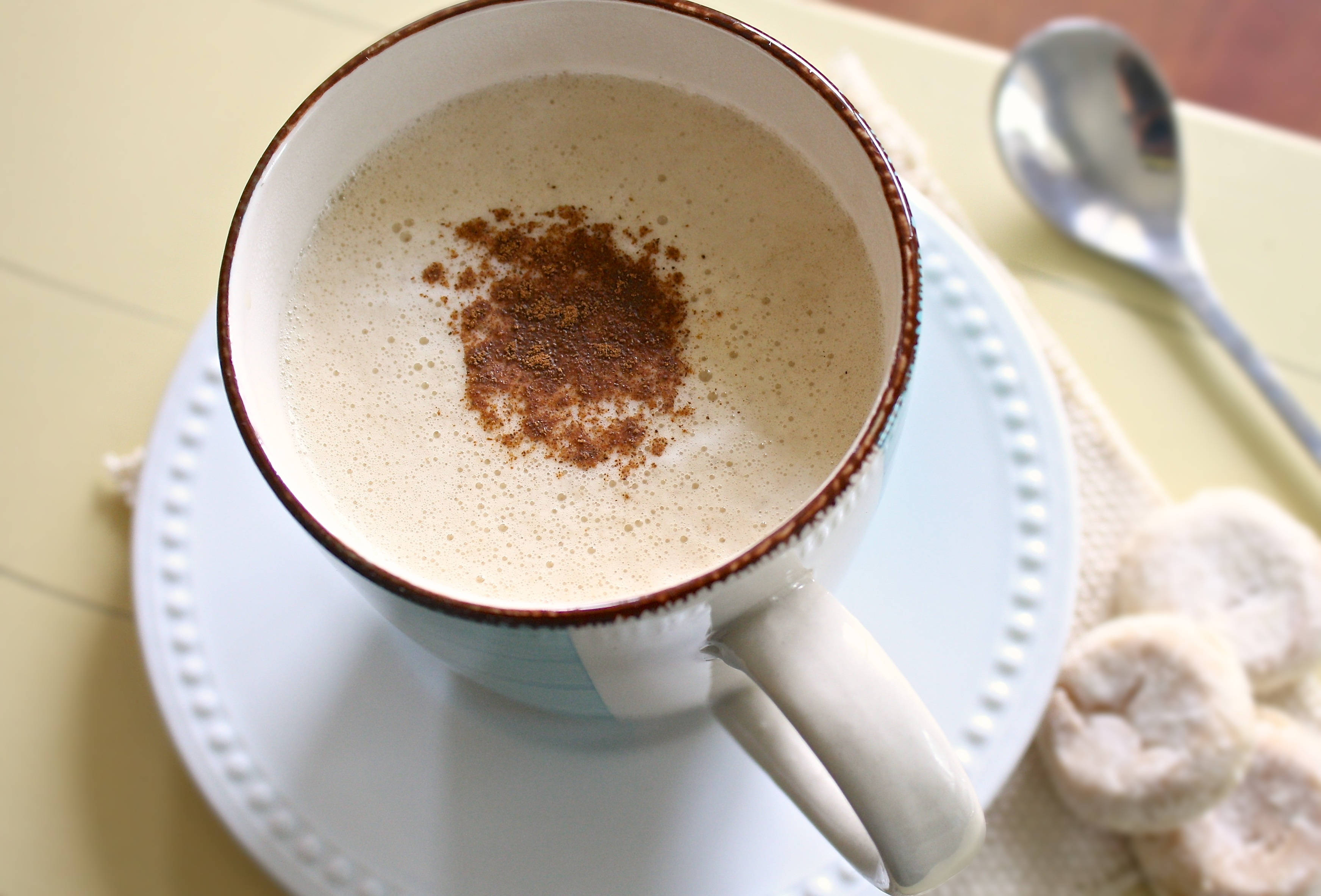 咖啡動圖 桌布 壁紙 — 一杯 creamy latte 撒上肉桂粉，放在陶瓷杯和白色碟子上，旁邊有糖餅乾和湯匙。