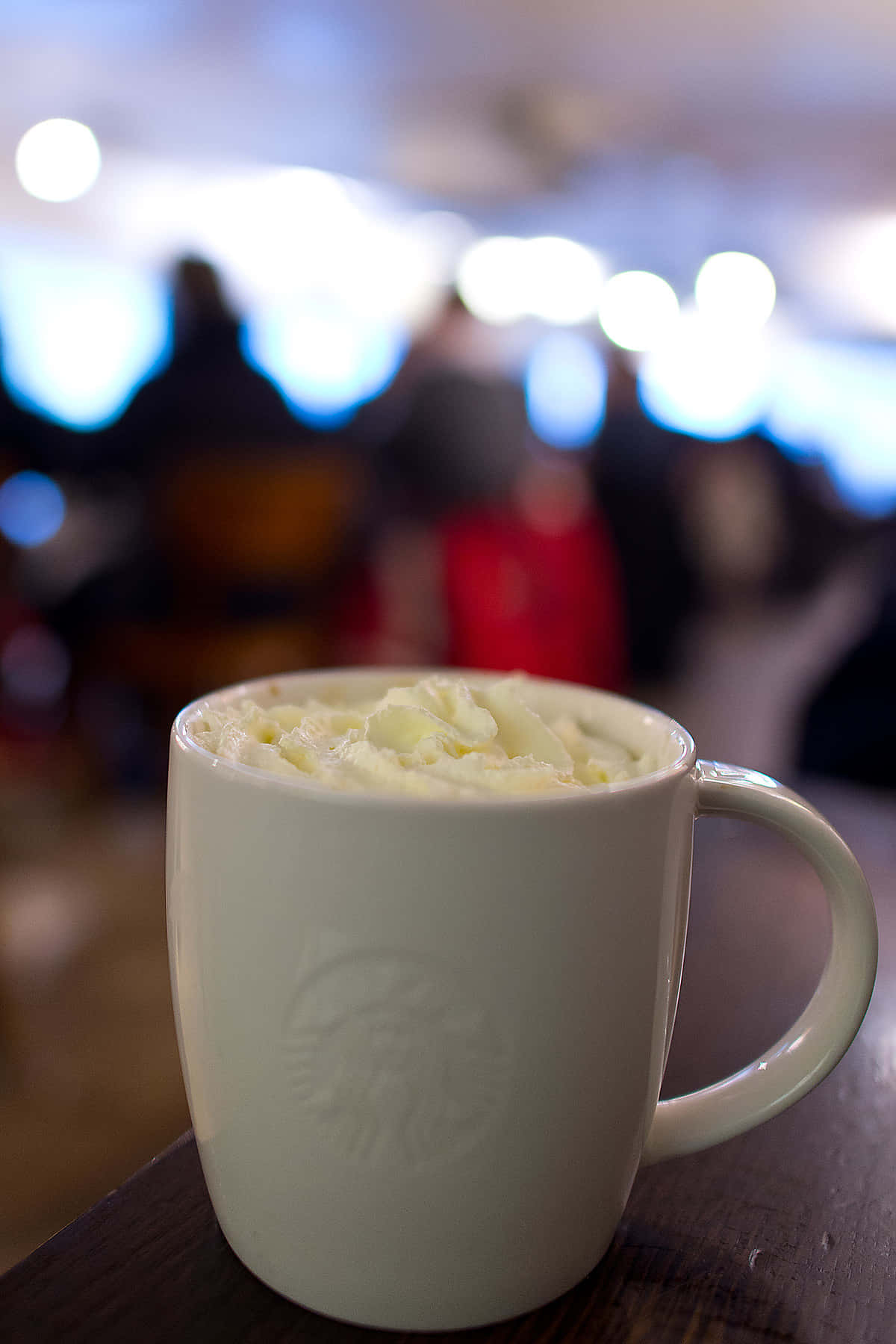 Winter Drinks 桌布 壁紙 — 一杯 creamy 泡沫咖啡杯放在木桌上，背景是模糊的咖啡店顧客和閃爍的燈光。