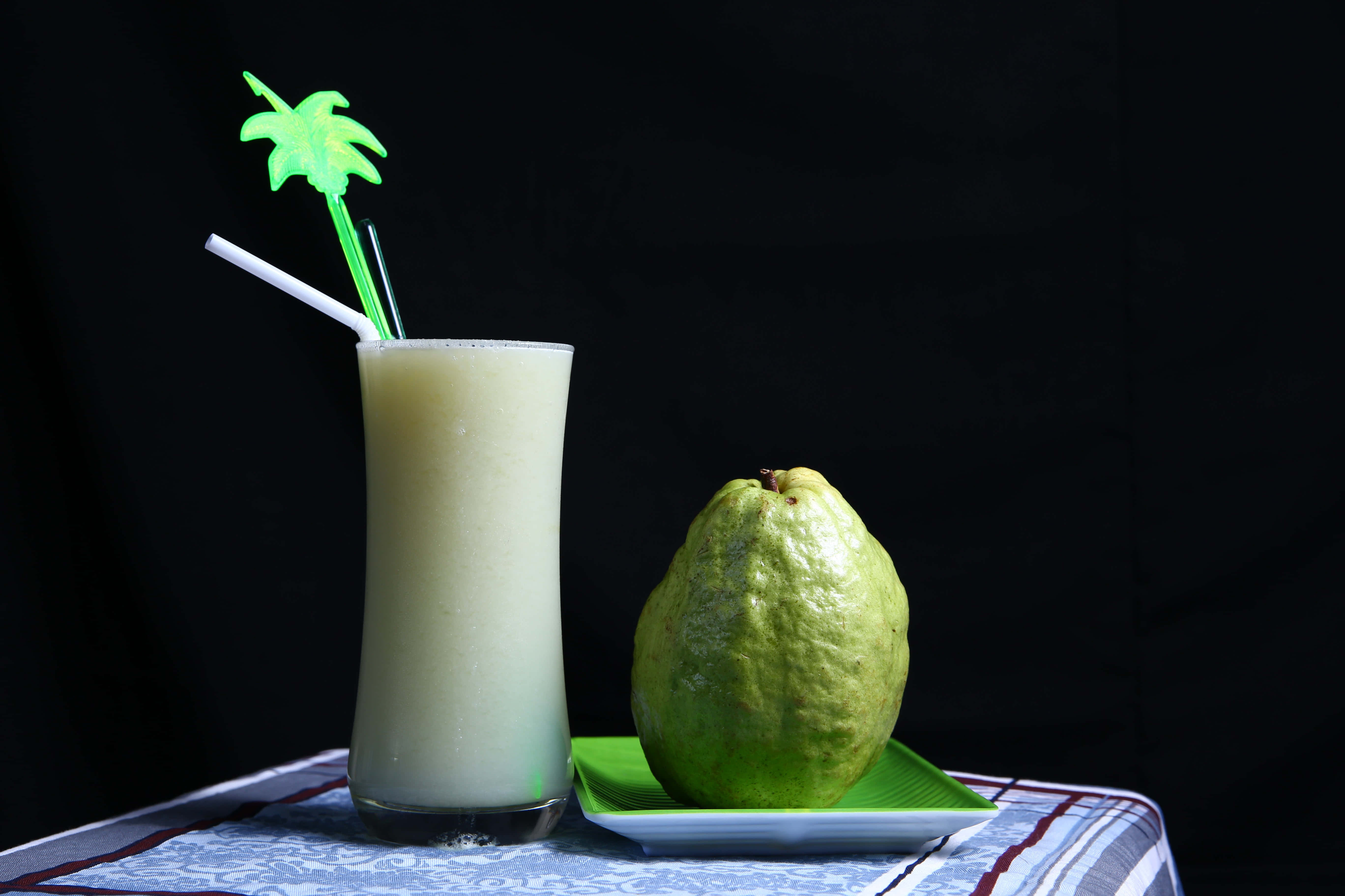 Summer Drinks 桌布 壁紙 — 一杯 creamy 淡綠色芭樂奶昔放在高腳玻璃杯中，配上綠色棕櫚吸管，旁邊的綠色盤子上放著一個成熟完整的芭樂，背景為黑色。