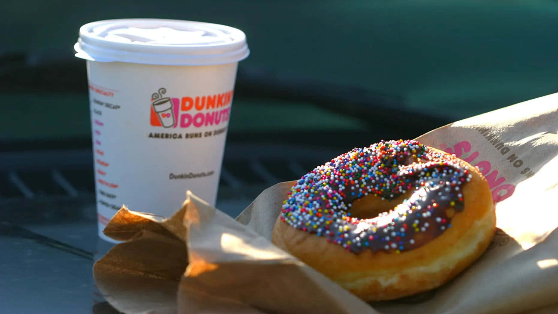 Dunkin Donuts 桌布 壁紙 — 一杯Dunkin' Donuts咖啡和一個巧克力碎甜甜圈放在溫暖陽光照射的深色表面上。