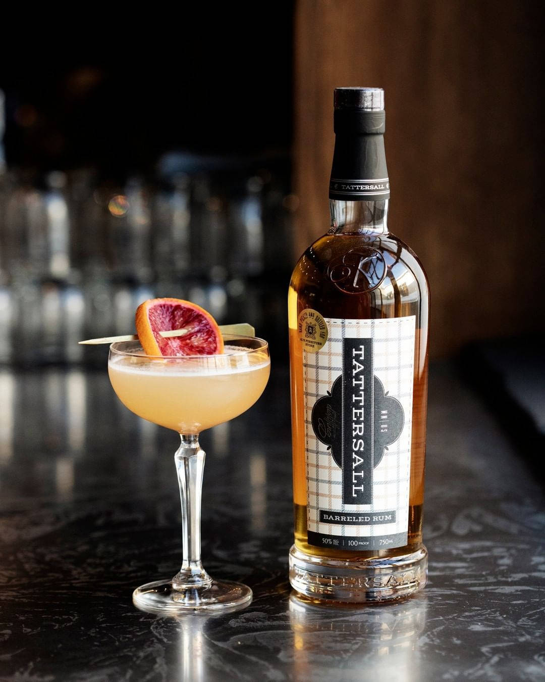 Drink 桌布 壁紙 — 一杯以血橙裝飾的金色雞尾酒，旁邊放著一瓶 Tattersall Barreled Rum，置於深色大理石吧檯上。