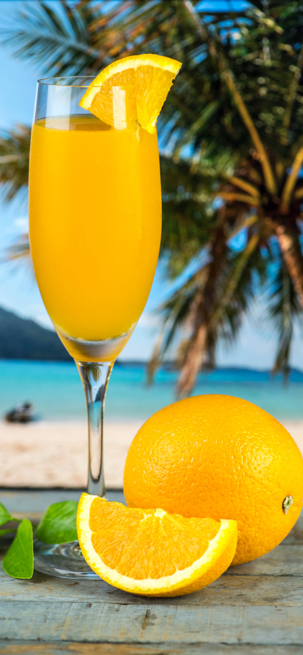 Tropical Drink 桌布 壁紙 — 一杯充滿活力的橙味雞尾酒盛載於笛形杯中，以一片橙裝飾，旁邊放著一個完整和已切開的橙，置於木桌上，背景是熱帶海灘和棕櫚樹。
