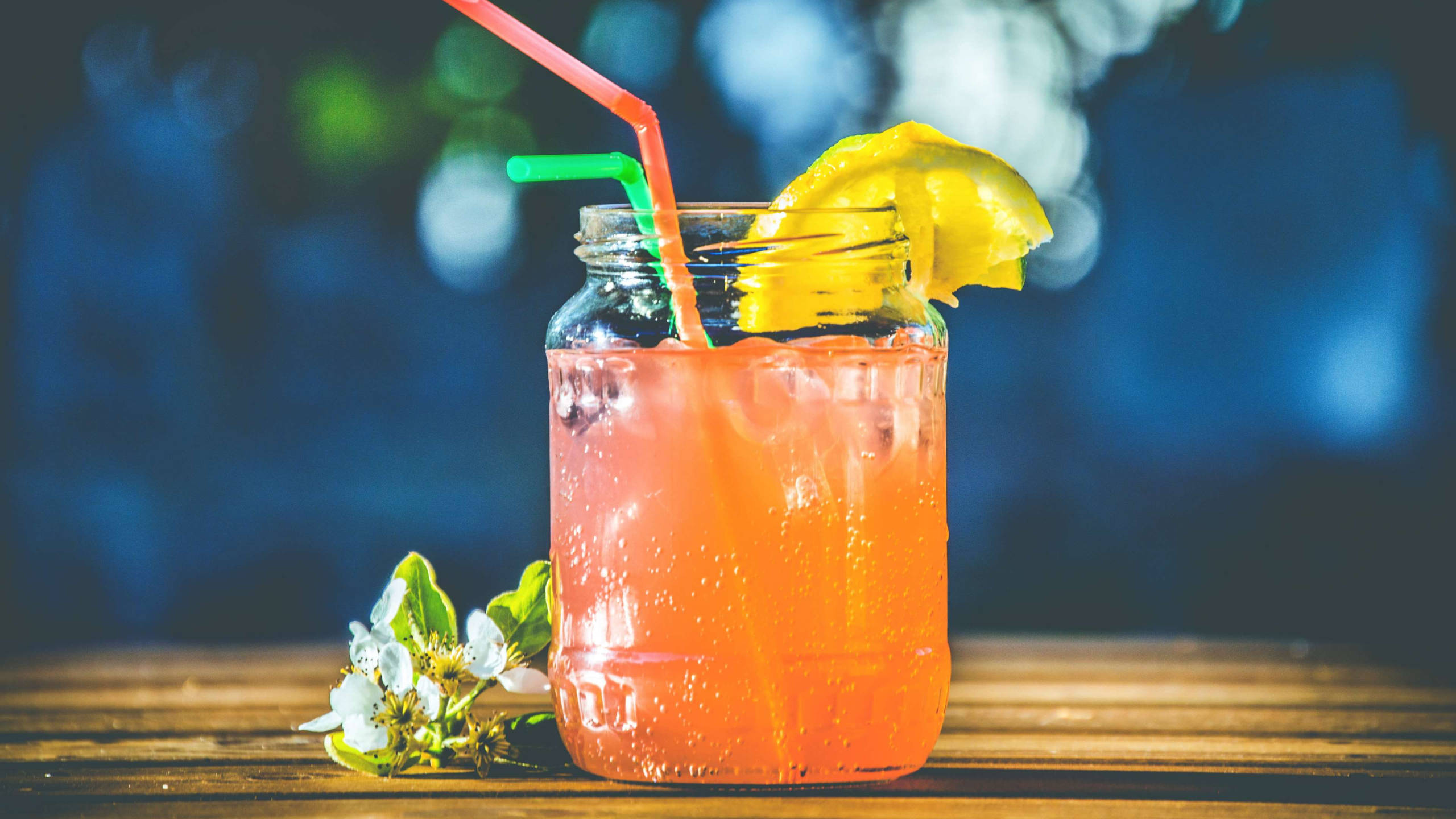 Tropical Drink 桌布 壁紙 — 一杯充滿活力的橙色冰飲，裝在梅森罐中，配上檸檬裝飾和雙吸管，放在木桌上，旁邊有白色花朵，背景是模糊的藍色。
