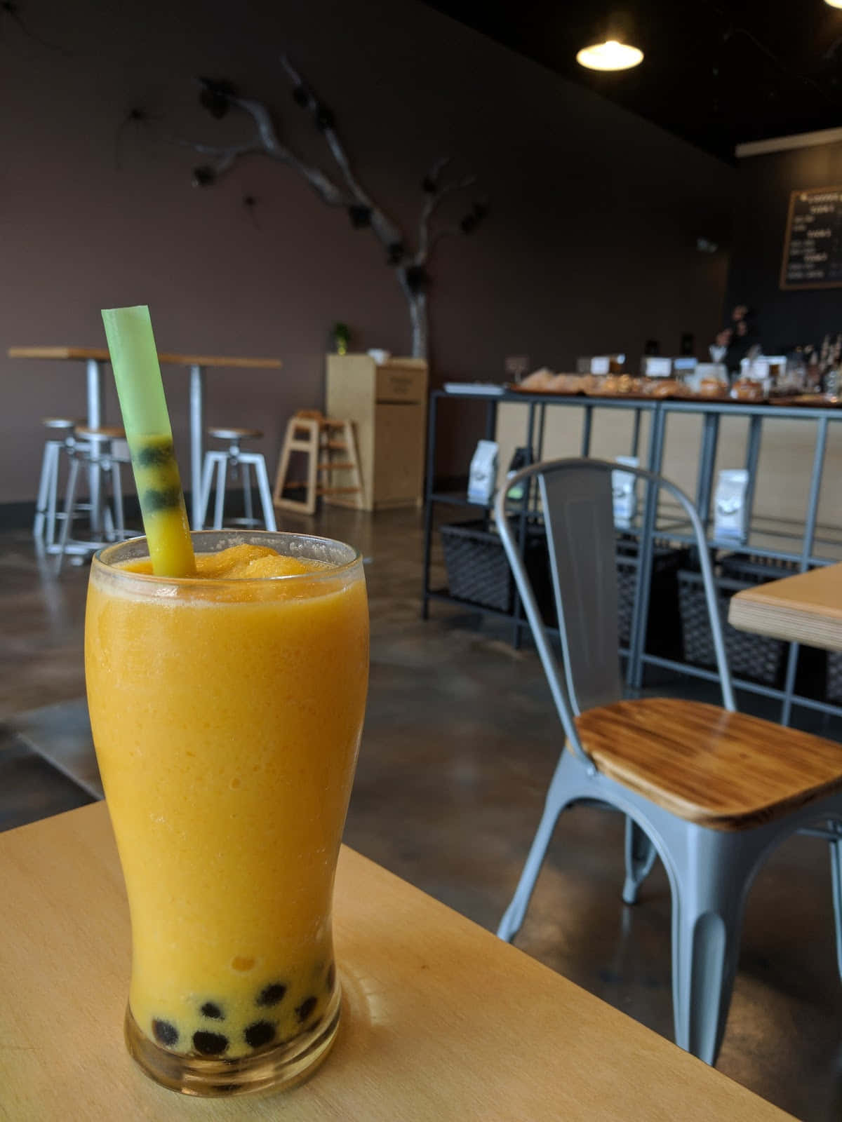 Boba Tea 桌布 壁紙 — 一杯充滿活力的橙色波霸飲料，搭配黑色珍珠和綠色吸管，放在一家舒適咖啡館的木桌上，周圍有金屬椅子和一棵光禿禿的樹雕塑。