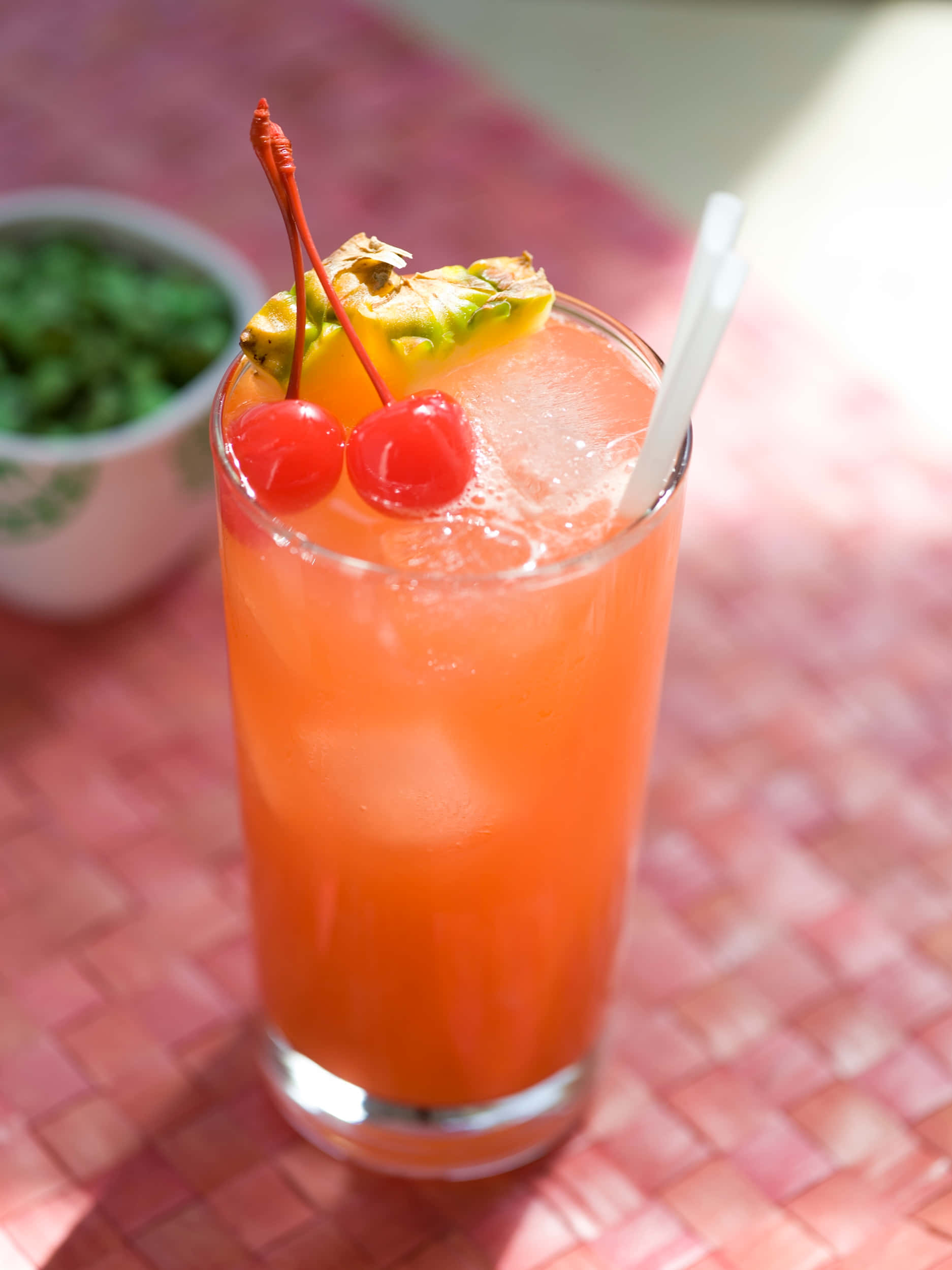 Winter Drinks 桌布 壁紙 — 一杯充滿活力的橙色熱帶雞尾酒，飾以鳳梨和櫻桃，放在編織的粉紅色墊子上，在明亮的陽光下。