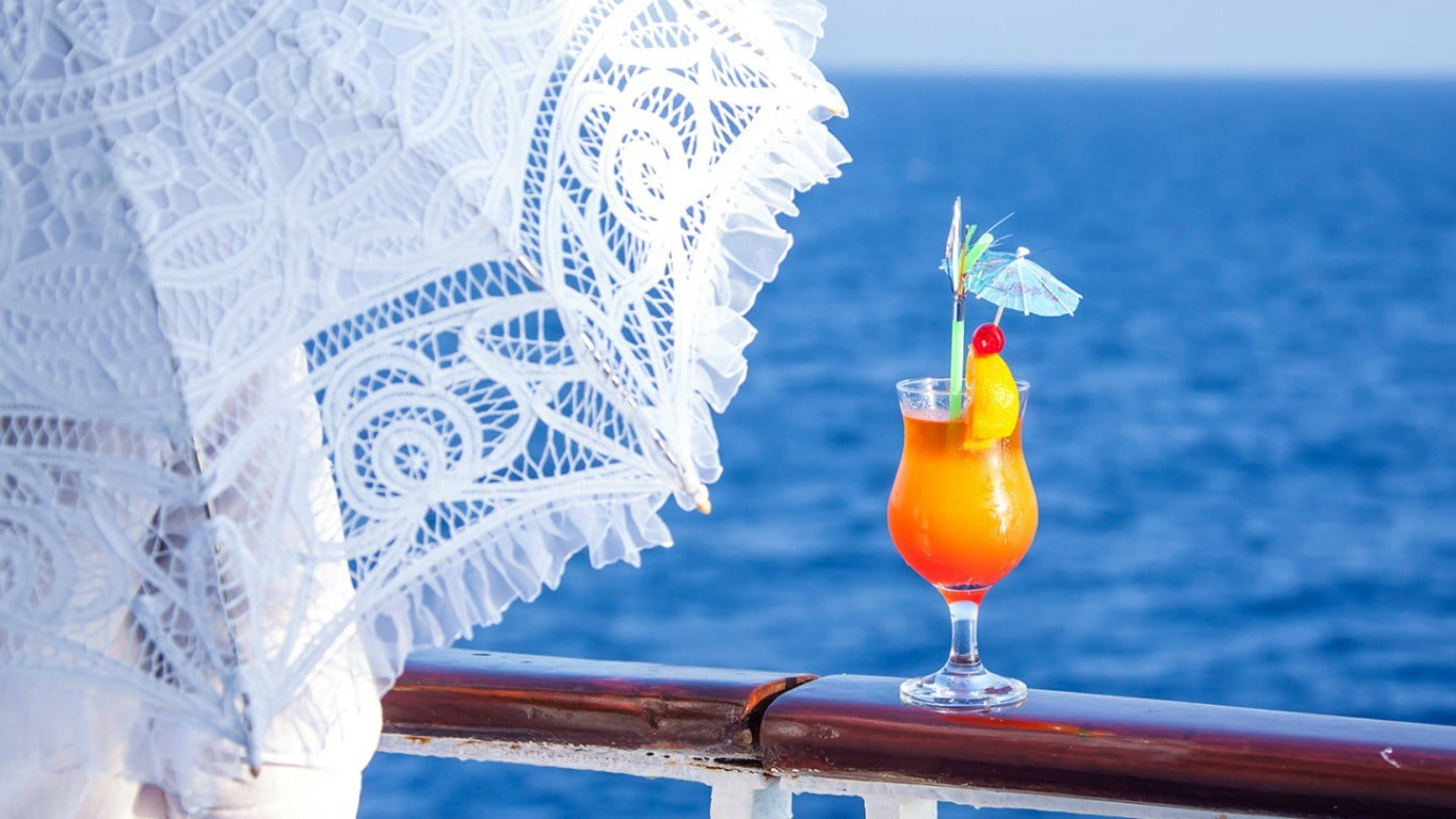 Tropical Drink 桌布 壁紙 — 一杯充滿活力的橙色熱帶飲品，配上櫻桃和傘飾，放在船欄杆上，旁邊有一把白色蕾絲陽傘，俯瞰深藍色的海洋。