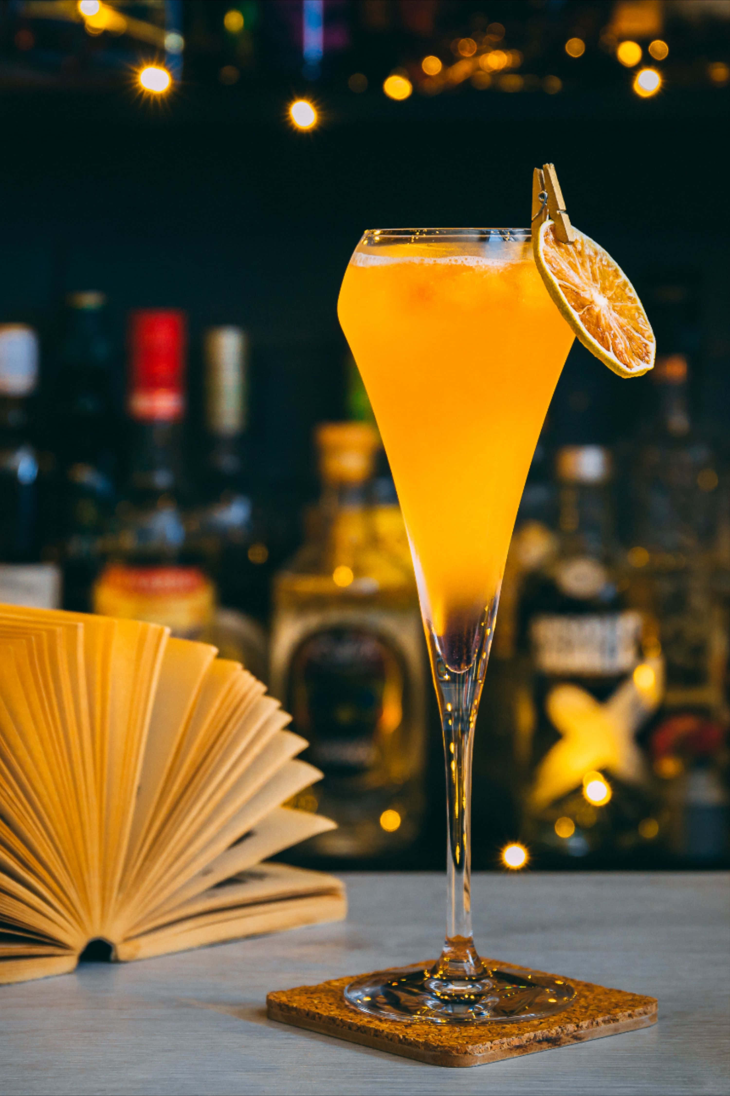 Winter Drinks 桌布 壁紙 — 一杯充滿活力的橙色雞尾酒，盛在高腳杯中，飾以一片柑橘，旁邊放著一本打開的書，背景是溫暖的散景燈光。