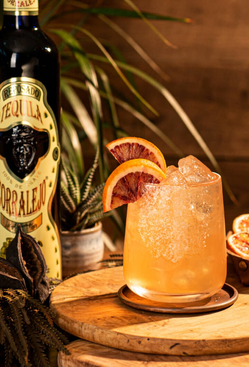 cocktail 桌布 壁紙 — 一杯充滿活力的橙色雞尾酒，飾以血橙片，旁邊放著一瓶雷波薩多龍舌蘭酒，置於質樸的木托盤上。