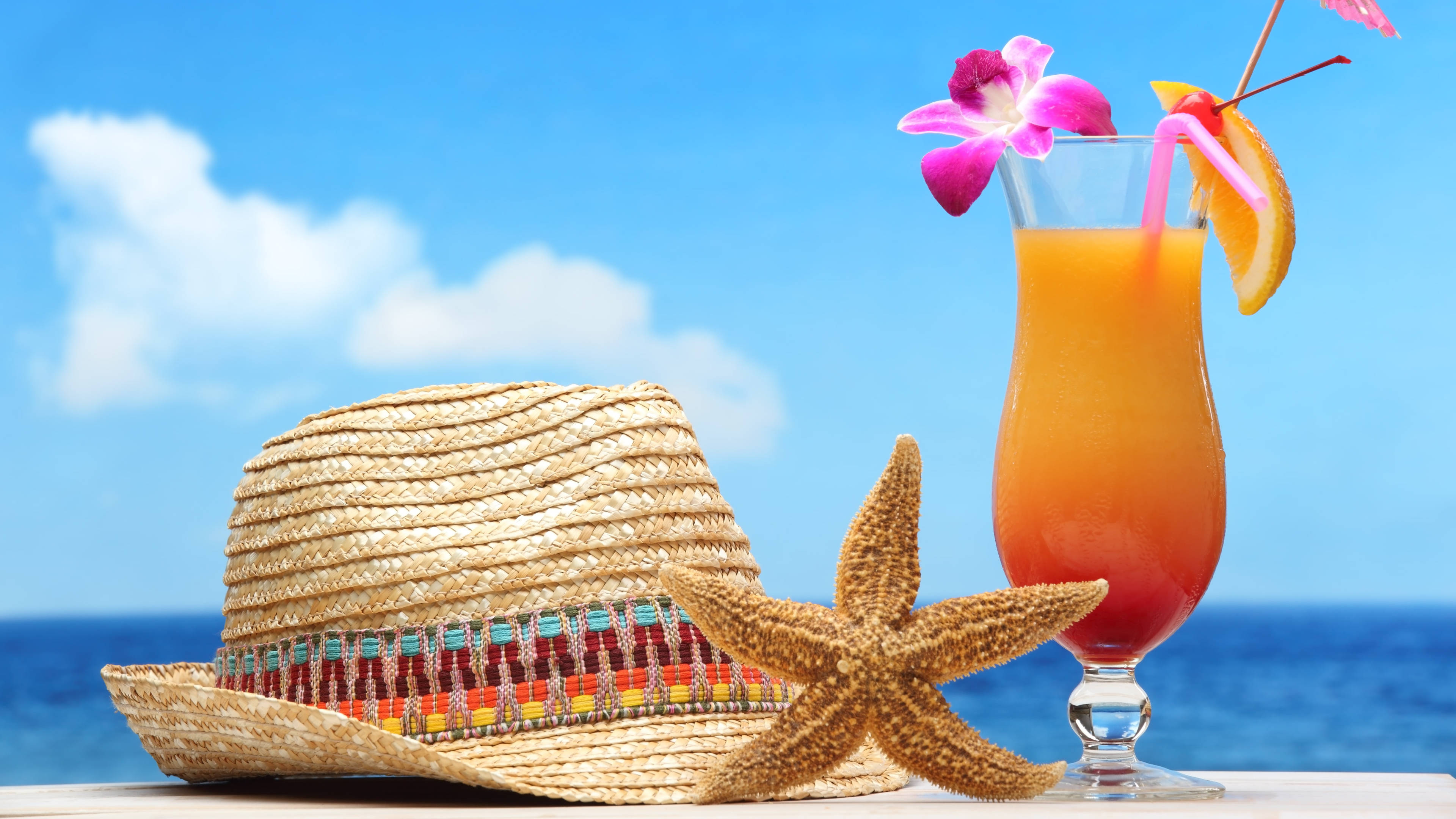 Tropical Drink 桌布 壁紙 — 一杯充滿活力的熱帶飲品，配以海星和草帽，放在桌子上，背景是明亮的藍色海洋和天空。