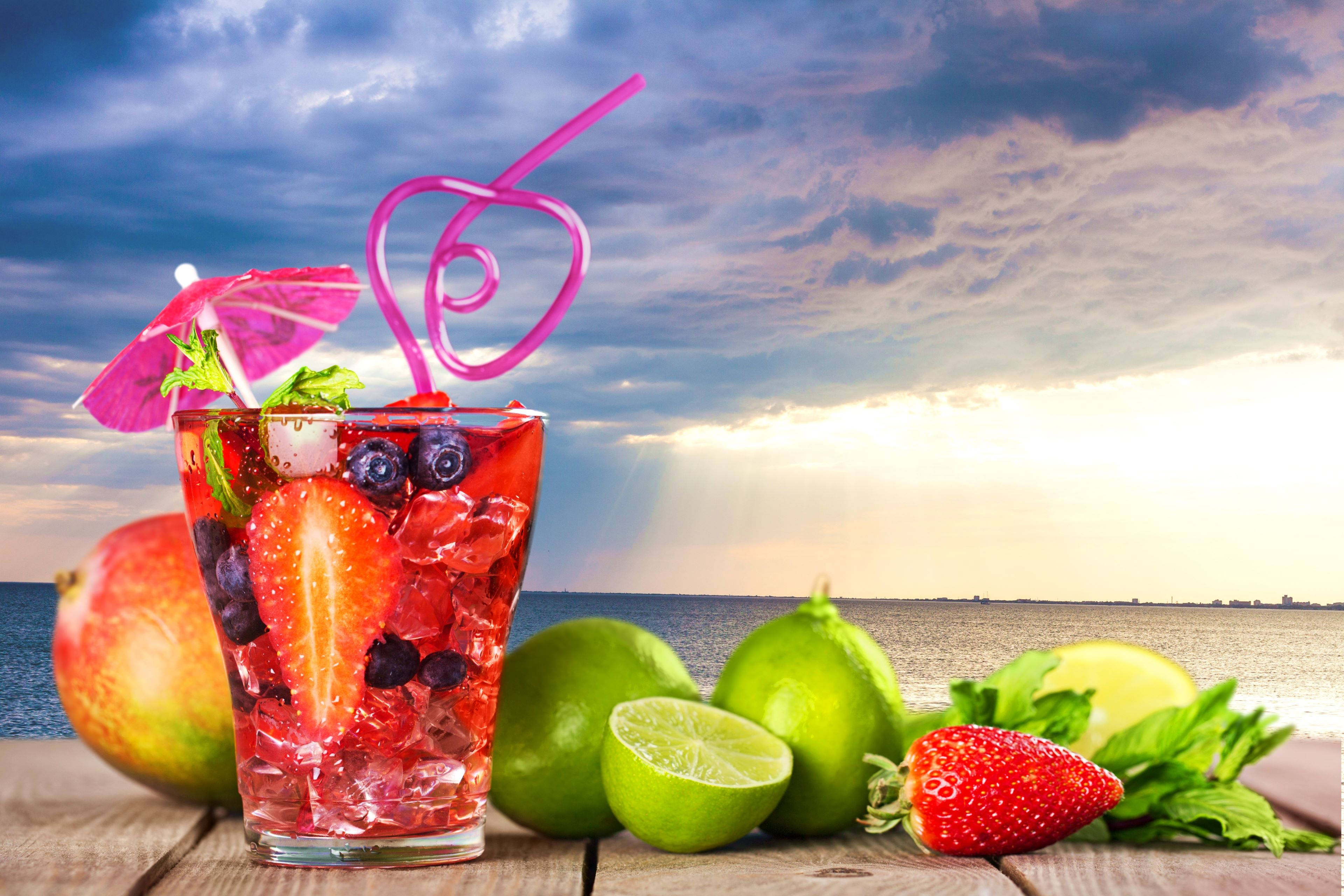 Tropical Drink 桌布 壁紙 — 一杯充滿活力的熱帶飲品，配有草莓、藍莓和薄荷，放在木桌上，旁邊有青檸、芒果和海洋上的日落。