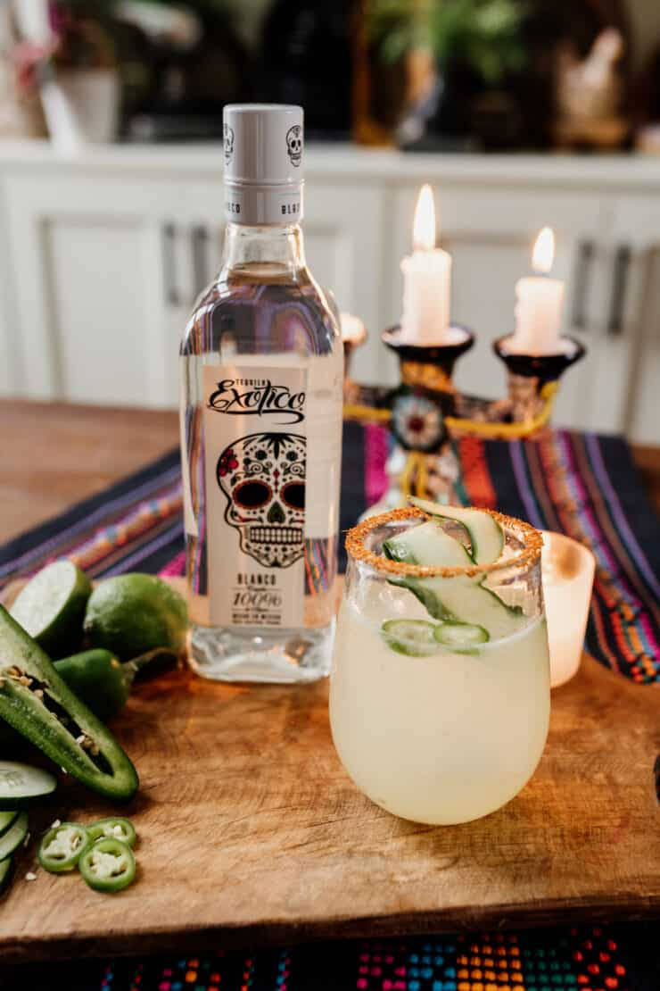 cocktail 桌布 壁紙 — 一杯充滿活力的瑪格麗特雞尾酒，配上墨西哥辣椒和萊姆裝飾，旁邊放著一瓶Exotico龍舌蘭酒，置於質樸的木板上，還有點燃的蠟燭和色彩繽紛的紡織品。
