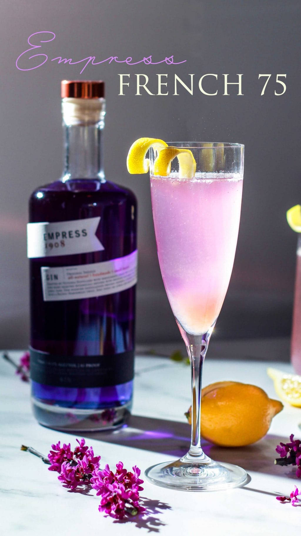 cocktail 桌布 壁紙 — 一杯充滿活力的粉紅色French 75雞尾酒，盛載於笛形杯中，以檸檬皮裝飾，旁邊放著一瓶紫色琴酒和新鮮花卉，置於大理石表面上。