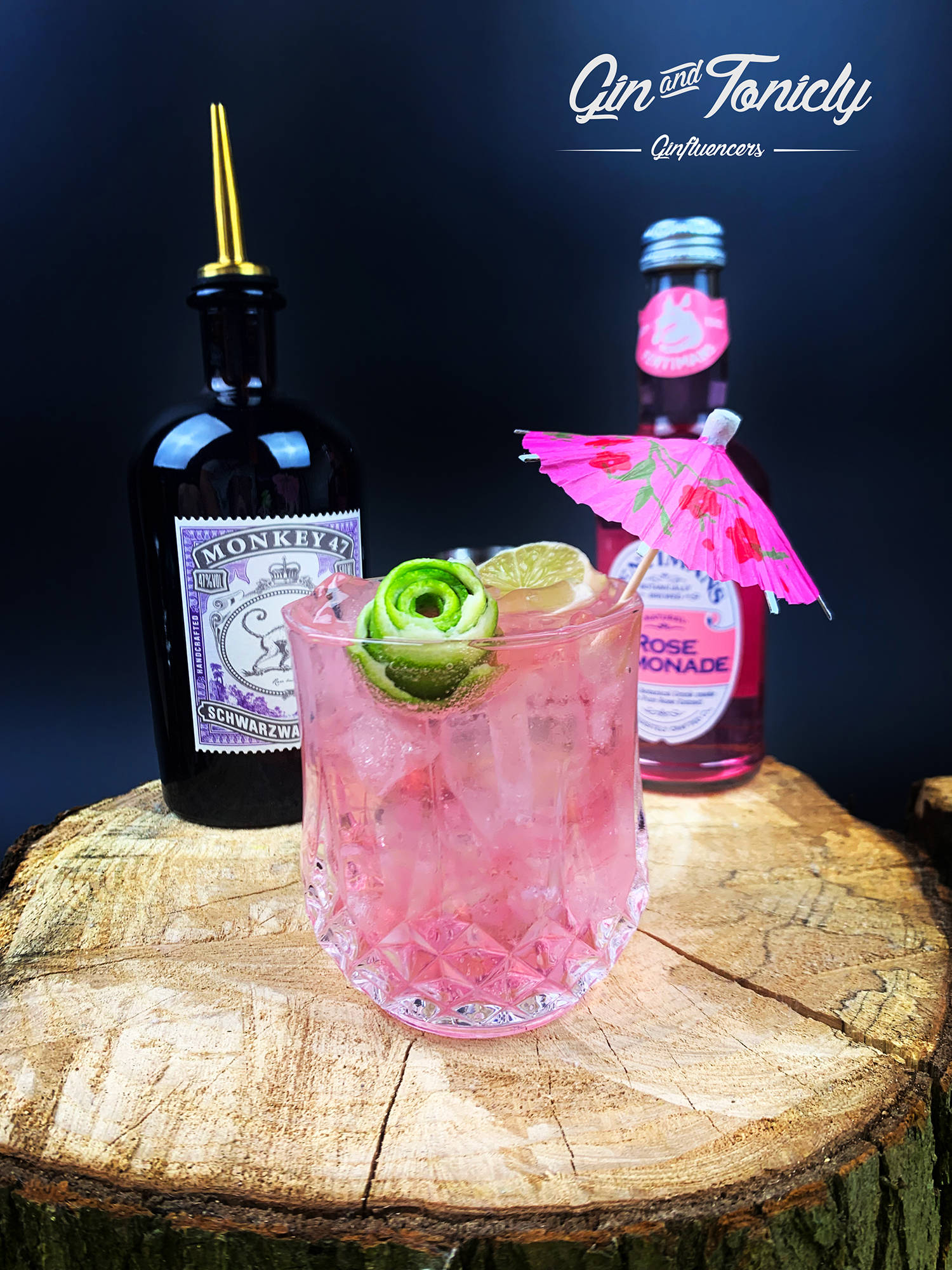 cocktail 桌布 壁紙 — 一杯充滿活力的粉紅色琴酒通寧雞尾酒，飾以萊姆和花卉圖案傘，放在質樸的木樁上，旁邊有一瓶Monkey 47琴酒和玫瑰檸檬水。