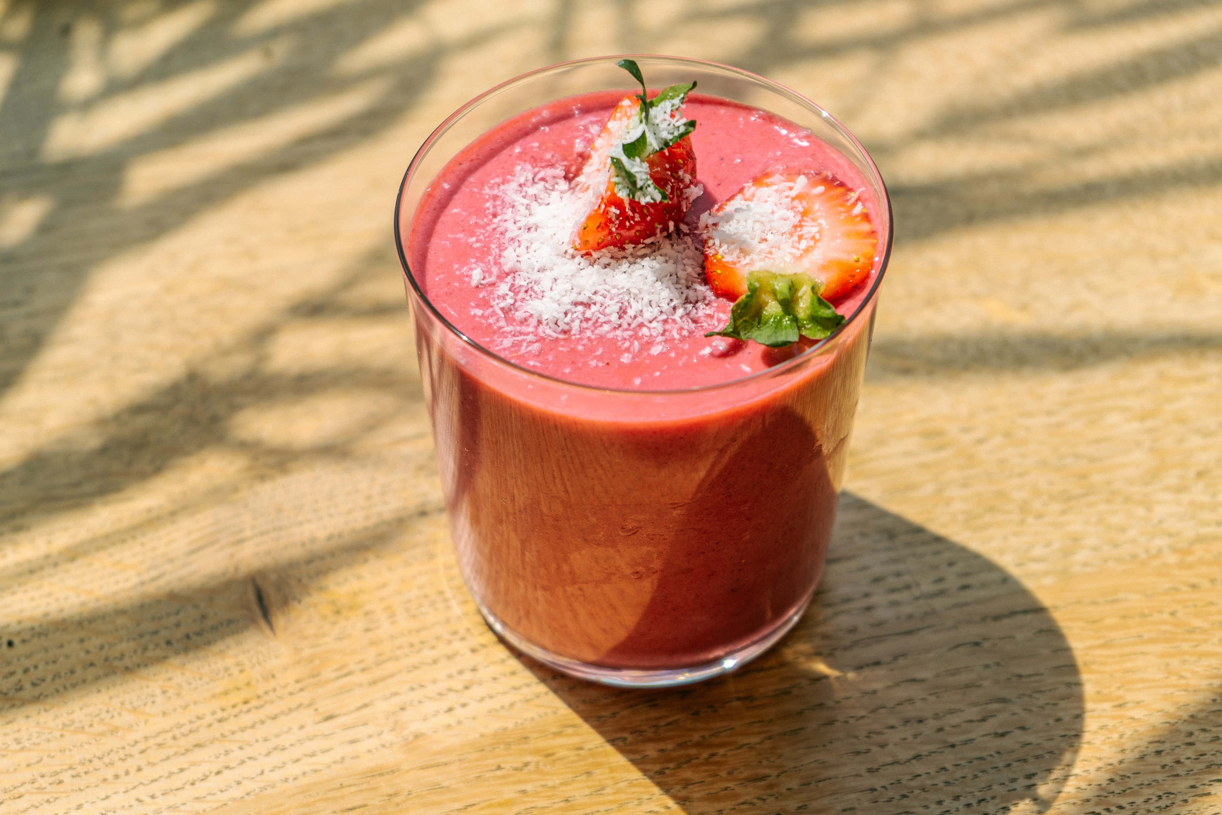 Summer Drinks 桌布 壁紙 — 一杯充滿活力的粉紅色草莓冰沙，上面撒上椰子片和新鮮草莓，放在陽光下的木桌上。