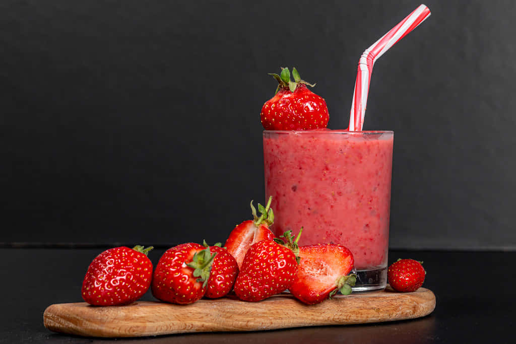 Strawberries 桌布 壁紙 — 一杯充滿活力的粉紅色草莓奶昔，放在木板上，配有條紋吸管和新鮮草莓，背景為深色。