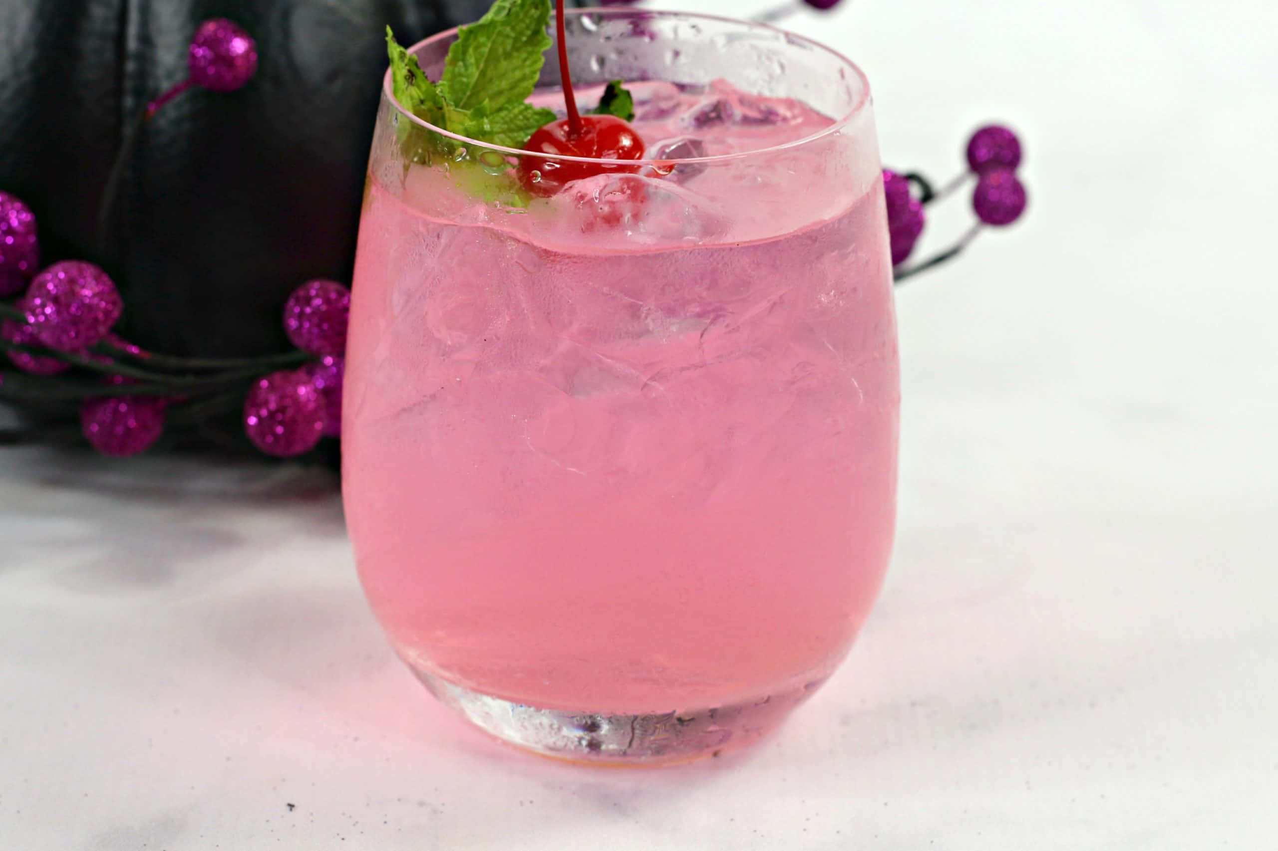Beautiful Drink 桌布 壁紙 — 一杯充滿活力的粉紅色雞尾酒，配上冰塊、櫻桃和薄荷裝飾，旁邊是閃閃發光的紫色莓果，放在白色表面上。