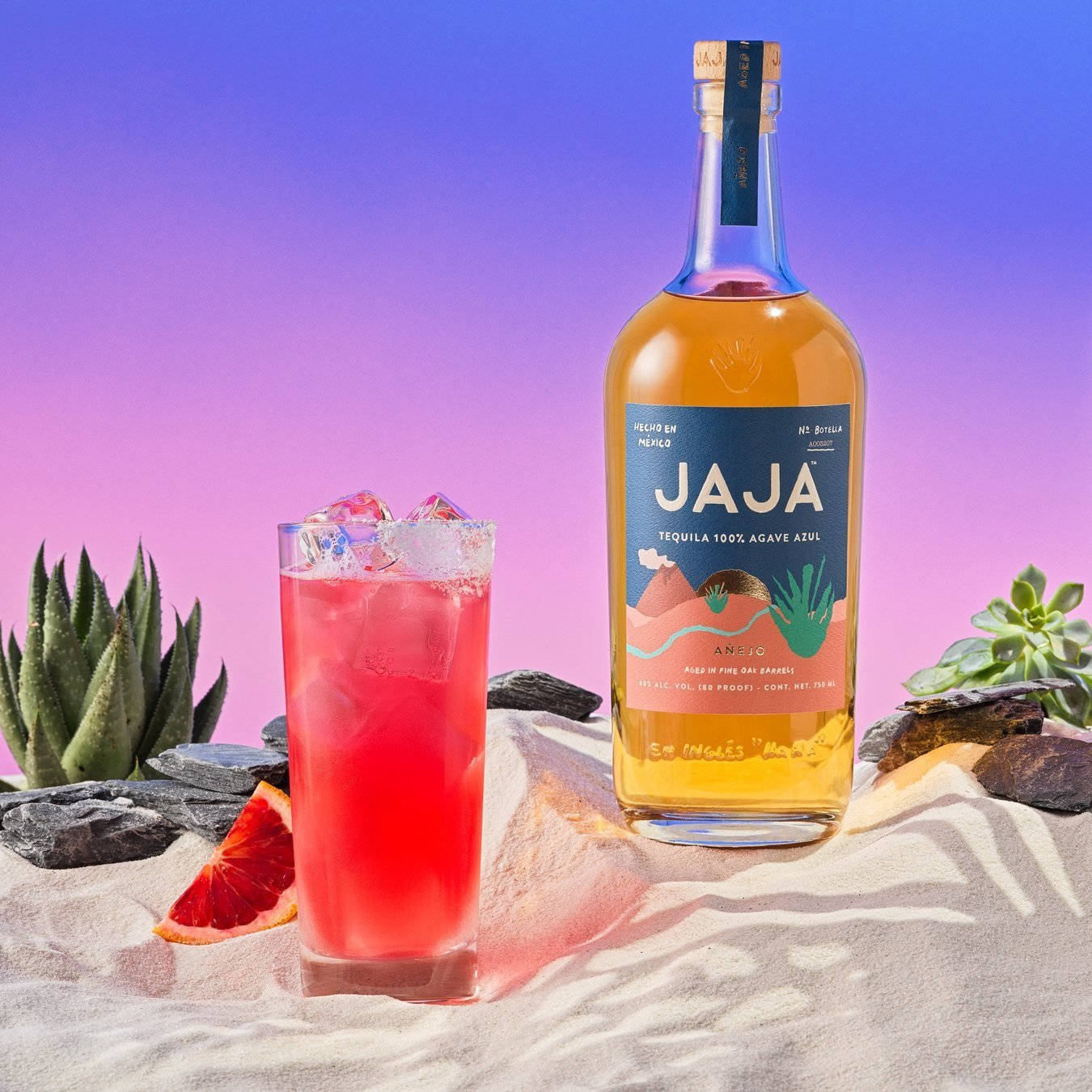 Tequila 桌布 壁紙 — 一杯充滿活力的粉紅色雞尾酒和一瓶Jaja龍舌蘭酒放在沙地上，旁邊有多肉植物，背景是漸變紫色天空。