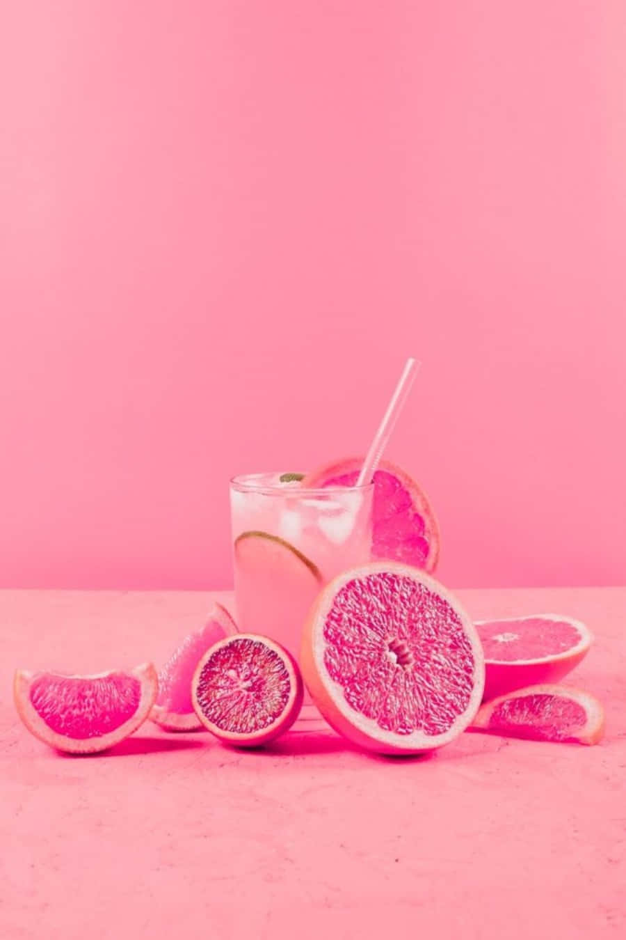 Pink Grapefruit 桌布 壁紙 — 一杯充滿活力的粉紅葡萄柚飲品，配有冰塊和吸管，周圍環繞著切片水果，放在相配的粉紅色表面上。