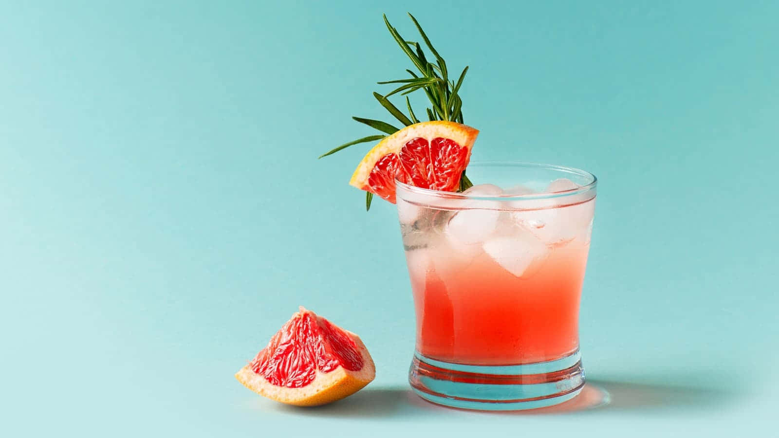 cocktail 桌布 壁紙 — 一杯充滿活力的粉紅西柚雞尾酒，配上冰塊和迷迭香裝飾，放在半個西柚旁邊，背景是柔和的青綠色。