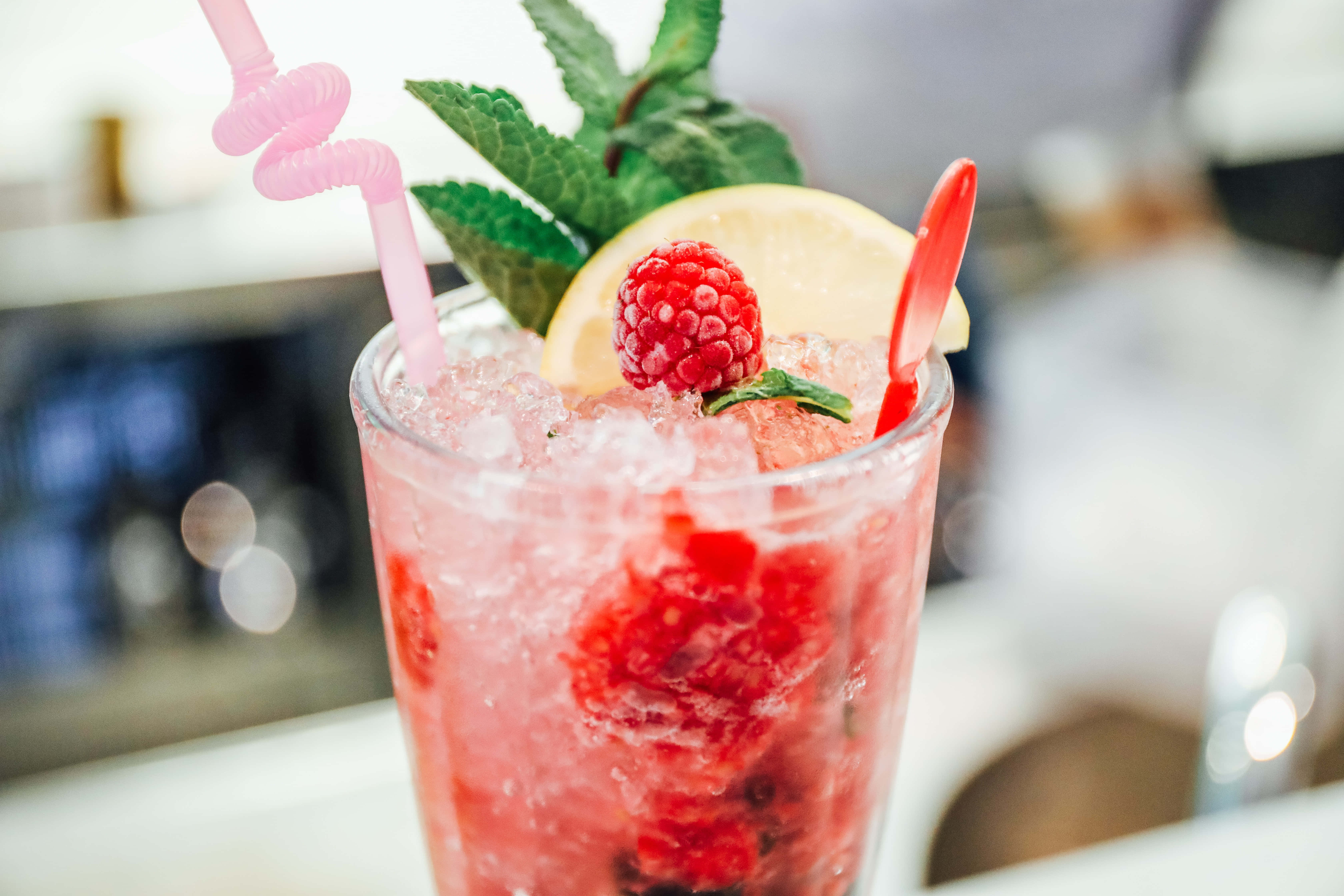 Summer Drinks 桌布 壁紙 — 一杯充滿活力的粉紅覆盆子雞尾酒，配上冰塊、薄荷、檸檬和彎曲吸管，放在酒吧檯上。