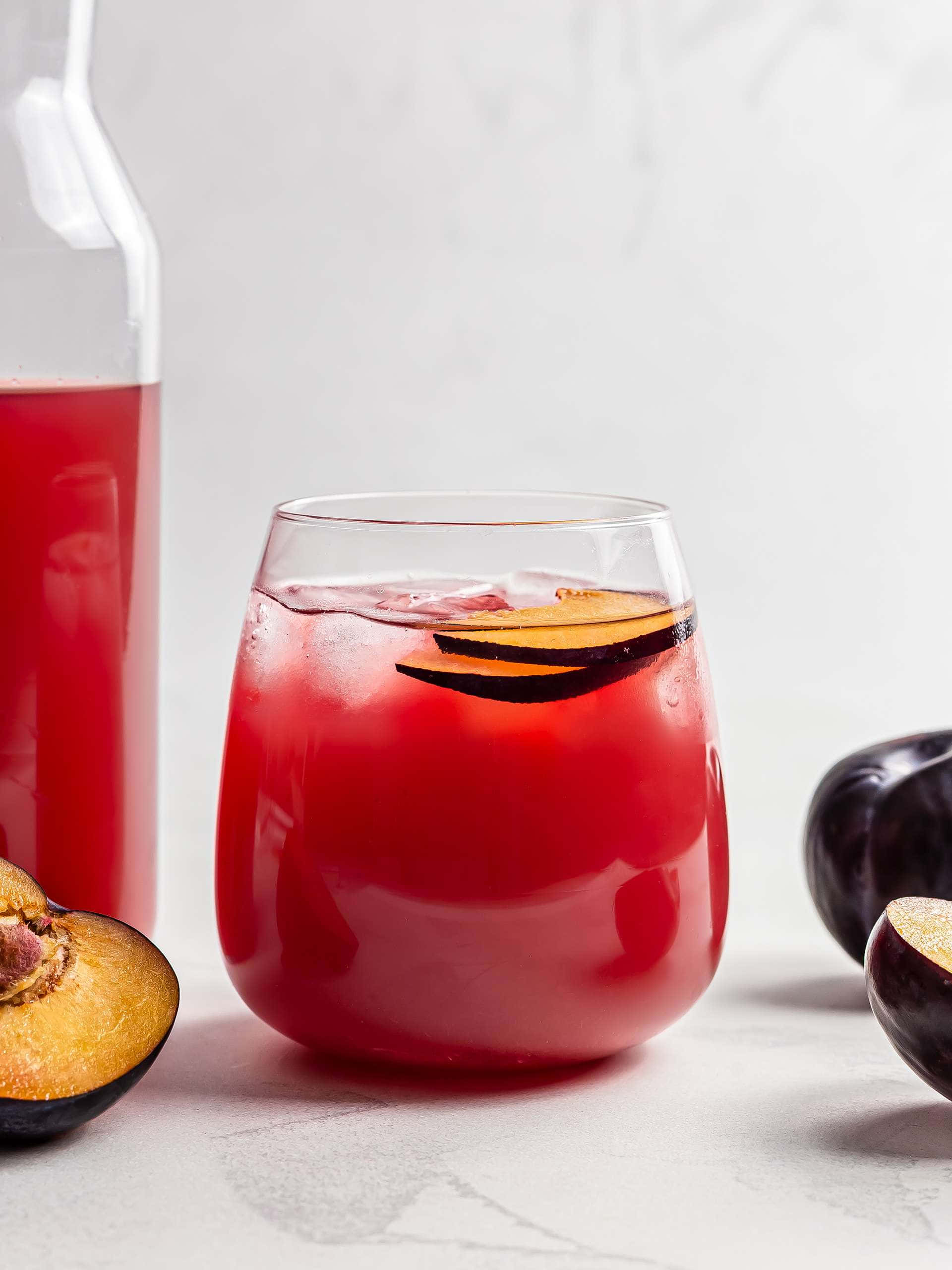 Red Plum 桌布 壁紙 — 一杯充滿活力的紅梅飲料，加冰和水果片，旁邊放著一瓶酒和切半的李子，置於大理石表面上。