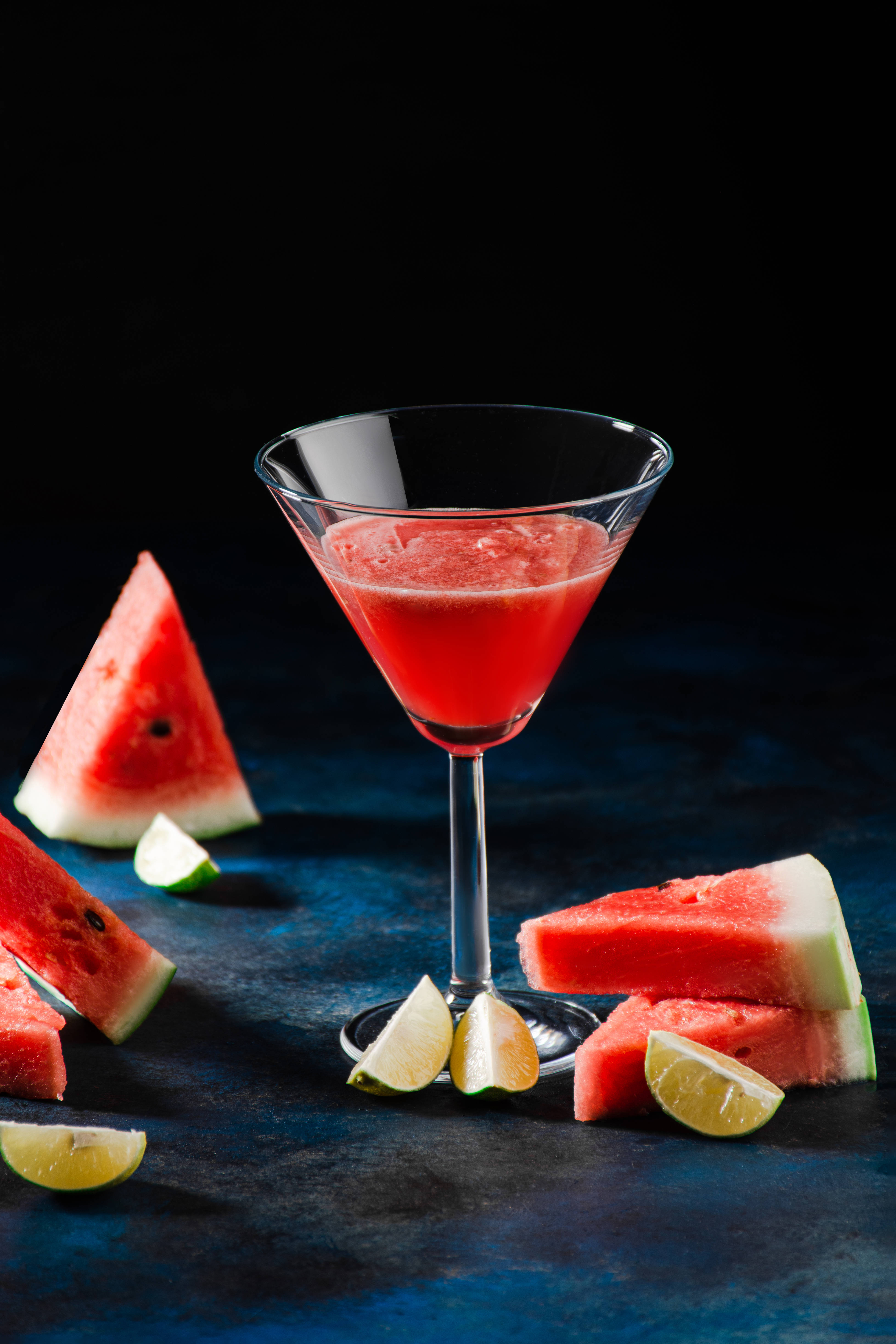 Watermelon 桌布 壁紙 — 一杯充滿活力的紅色西瓜雞尾酒，盛在馬丁尼杯中，周圍是新鮮西瓜片和萊姆角，置於深藍色紋理表面上。