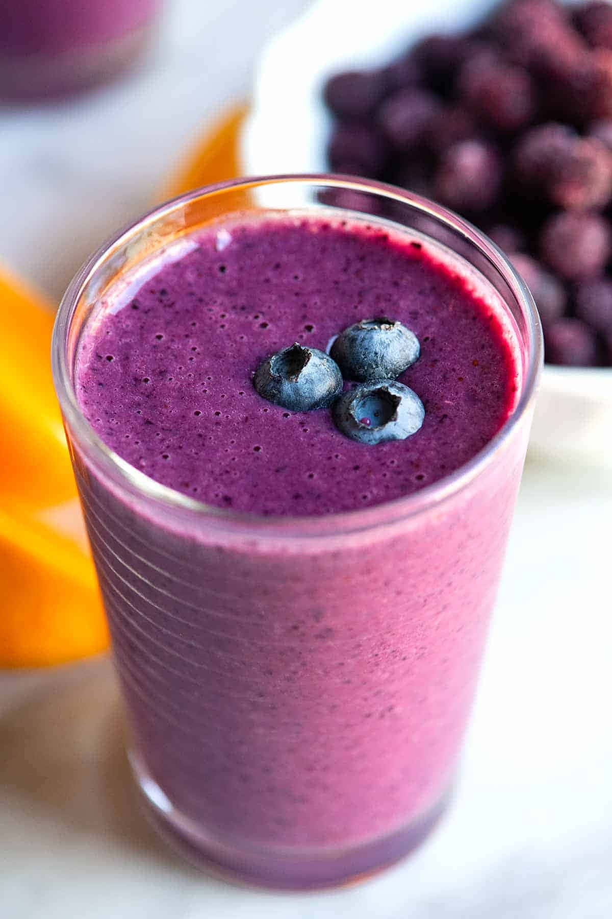 Blueberry Smoothie 桌布 壁紙 — 一杯充滿活力的紫色冰沙，上面放著新鮮藍莓，旁邊有一堆藍莓和柳橙片。