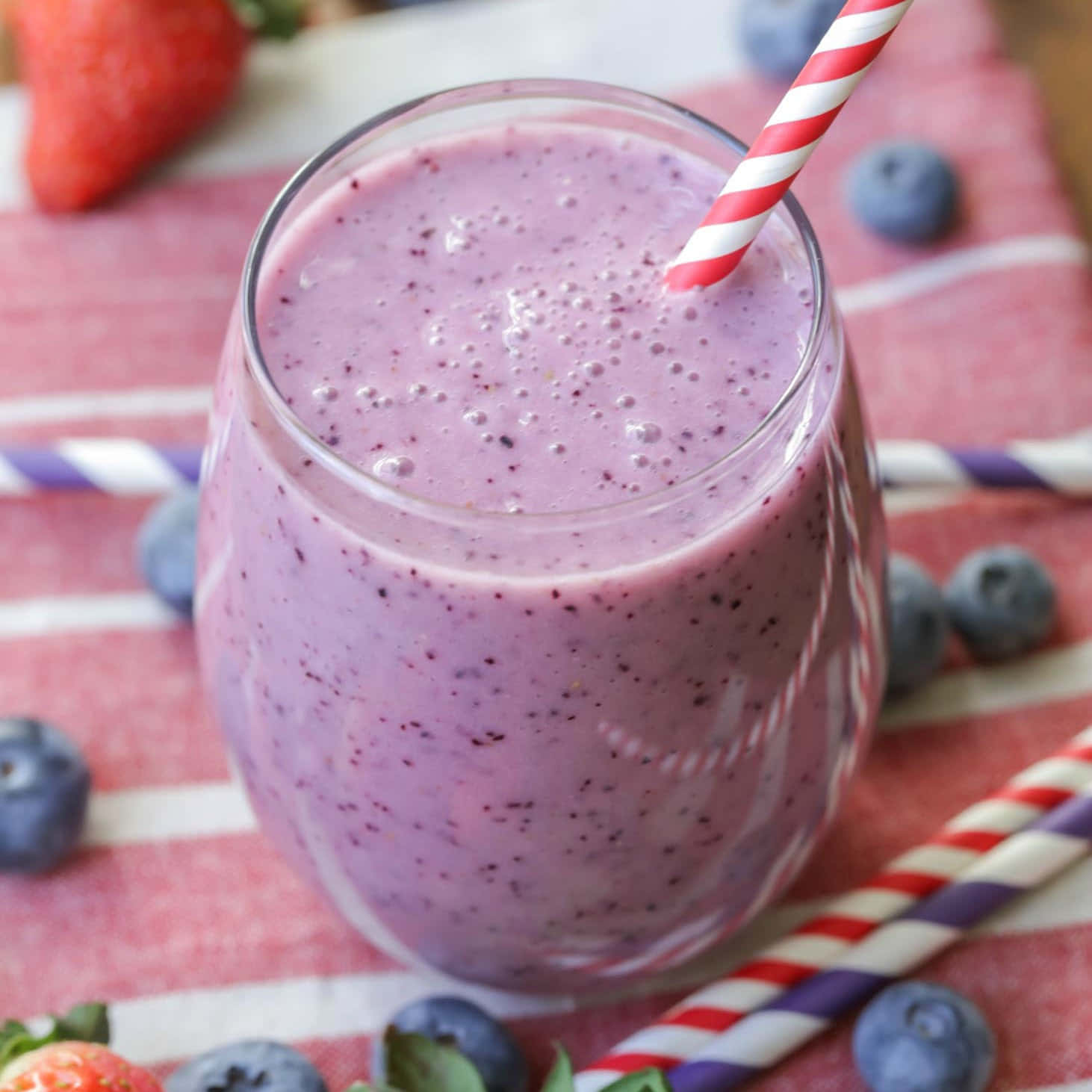 Blueberry Smoothie 桌布 壁紙 — 一杯充滿活力的紫色冰沙，放在玻璃杯中，插著紅白條紋吸管，周圍是新鮮藍莓和草莓，置於條紋布上。
