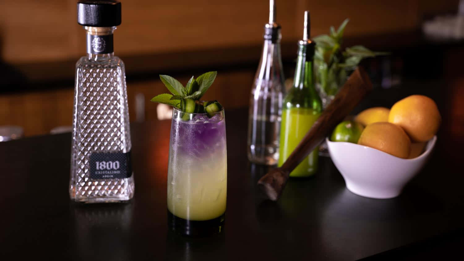 alcohol 桌布 壁紙 — 一杯充滿活力的紫色和黃色雞尾酒，飾以薄荷，旁邊放著一瓶1800 Crystaline龍舌蘭酒和柑橘類水果，在昏暗的吧檯上。