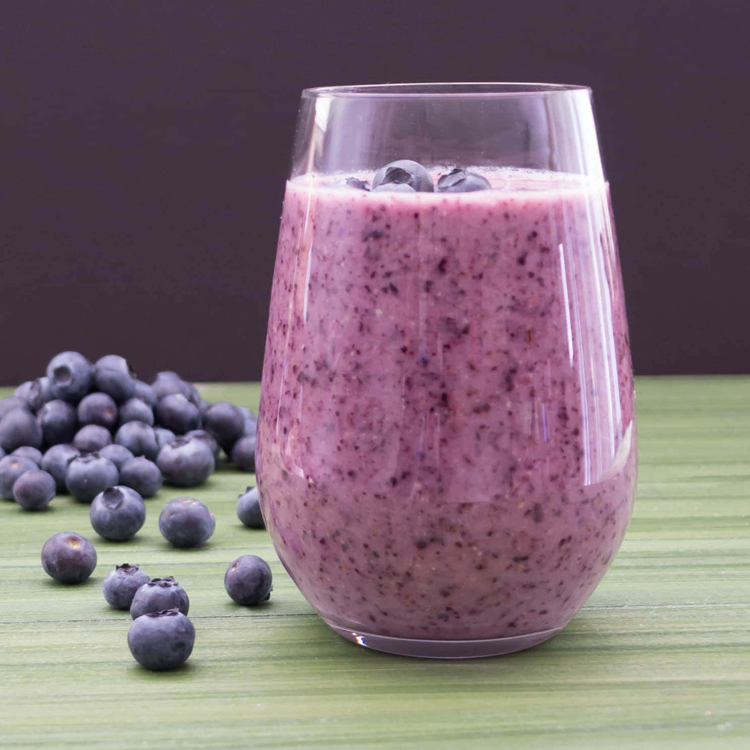 Blueberry Smoothie 桌布 壁紙 — 一杯充滿活力的紫色藍莓冰沙，上面放著新鮮藍莓，旁邊有一堆完整的藍莓放在綠色木質表面上。