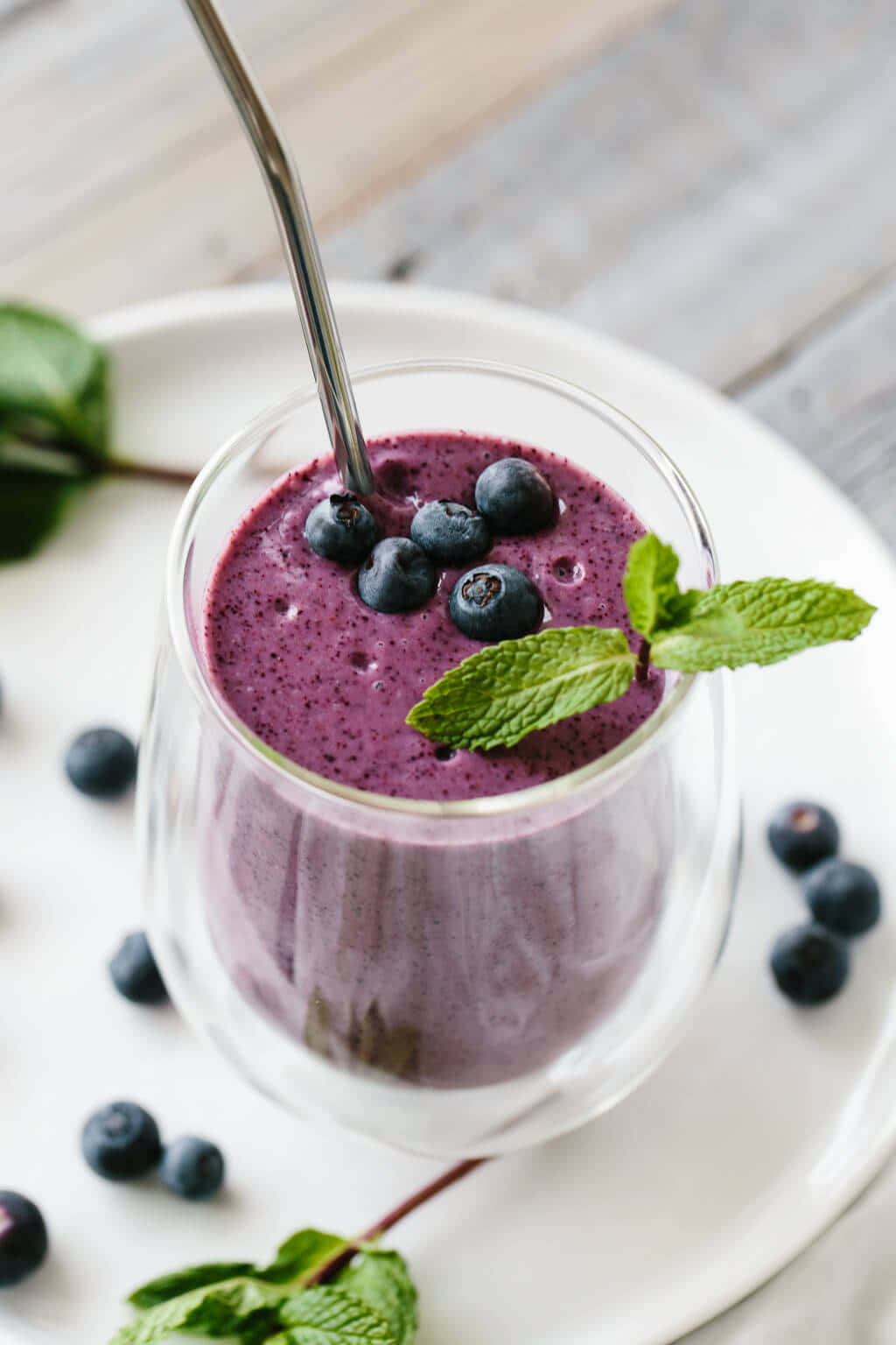 Blueberry Smoothie 桌布 壁紙 — 一杯充滿活力的紫色藍莓冰沙，上面放上新鮮藍莓和薄荷葉，置於白色盤子上。