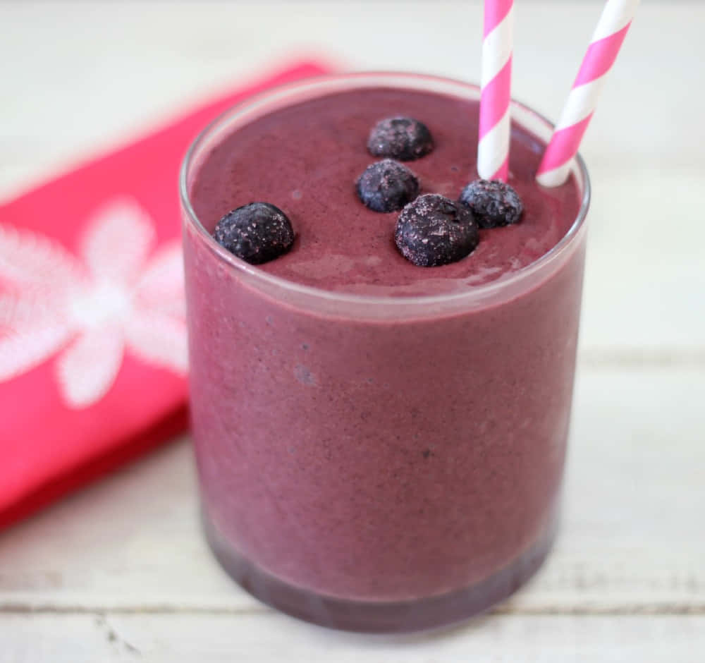 Blueberry Smoothie 桌布 壁紙 — 一杯充滿活力的紫色藍莓奶昔，上面放上新鮮莓果和粉紅條紋吸管，放在白色木質表面上，旁邊有一張紅色花卉圖案餐巾。