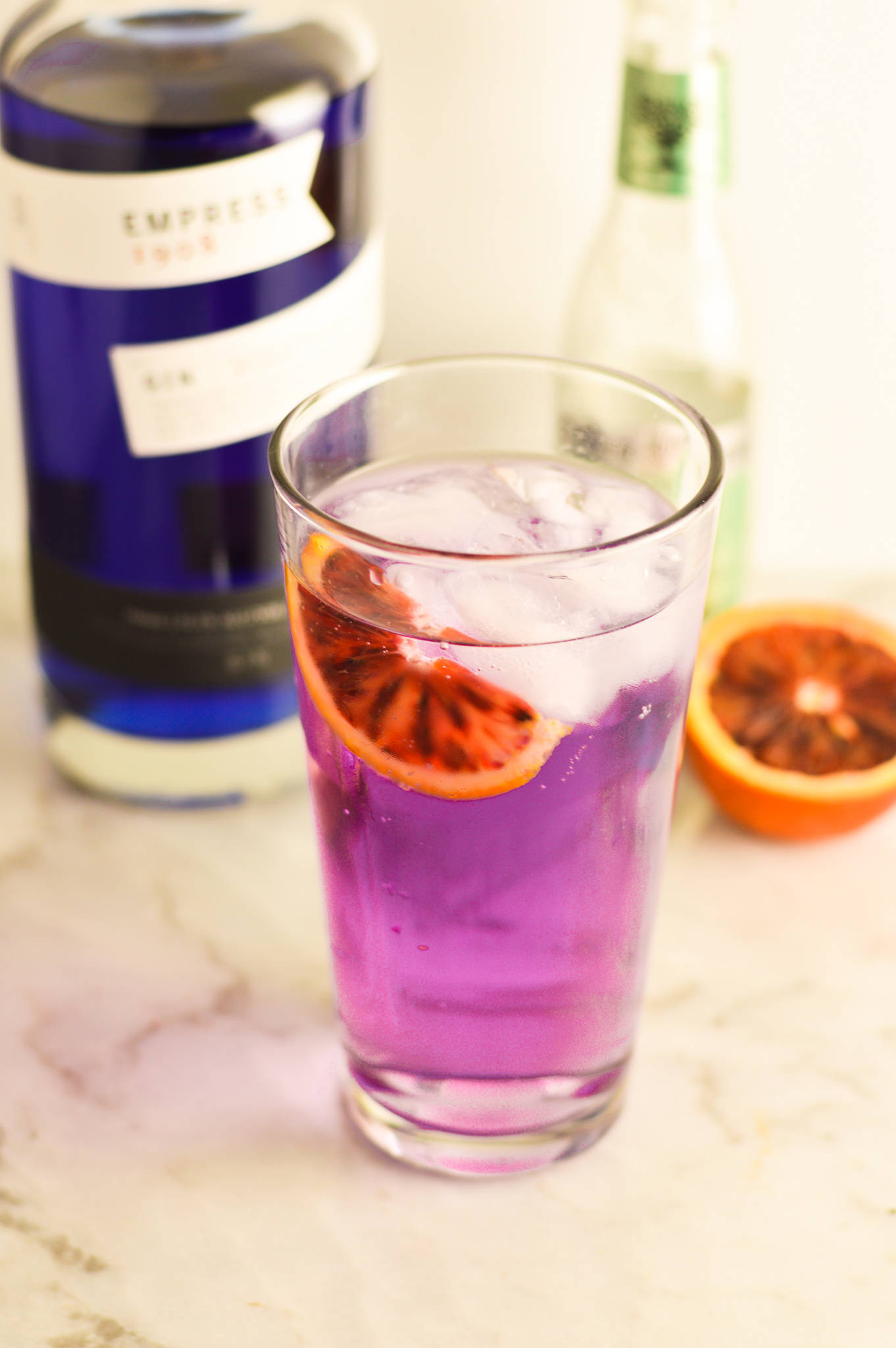 cocktail 桌布 壁紙 — 一杯充滿活力的紫色雞尾酒，加冰和血橙片，放在大理石檯面上，旁邊有一個藍色瓶子和切片橙。