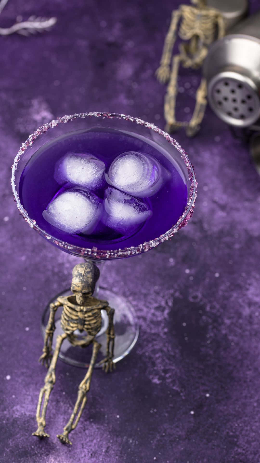 Spooky Cocktails 桌布 壁紙 — 一杯充滿活力的紫色雞尾酒，帶有亮粉邊緣和冰塊，放在一個由小型骷髏公仔手持的馬丁尼杯中，置於相配的紫色紋理表面上。
