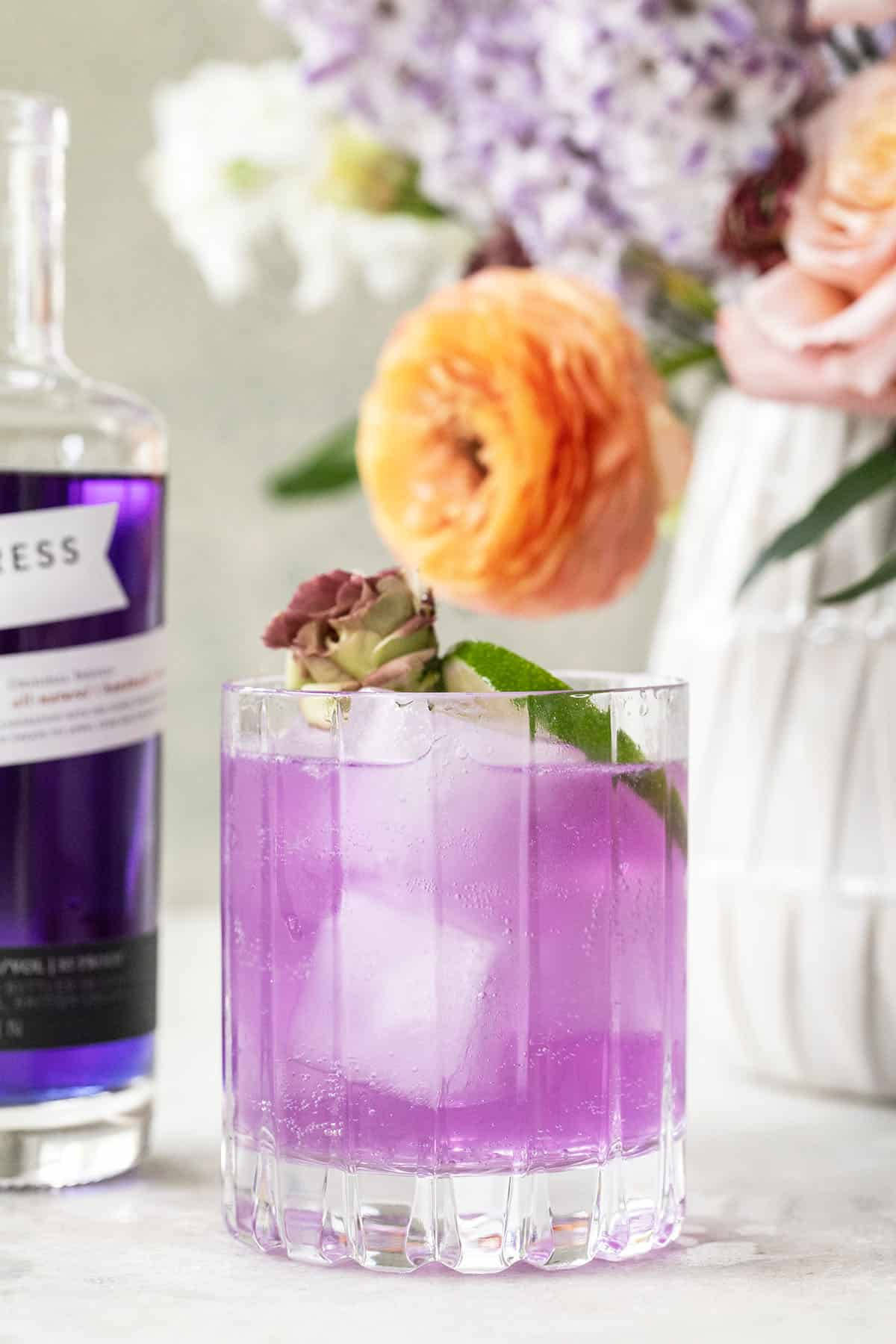cocktail 桌布 壁紙 — 一杯充滿活力的紫色雞尾酒，配上冰塊和花卉裝飾，旁邊放著一瓶酒和一束柔和粉彩花。