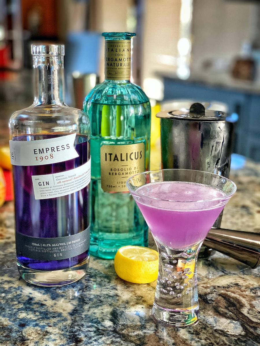 cocktail 桌布 壁紙 — 一杯充滿活力的紫色雞尾酒放在馬丁尼杯中，旁邊是Empress Gin和Italicus利口酒瓶，置於斑點檯面上。