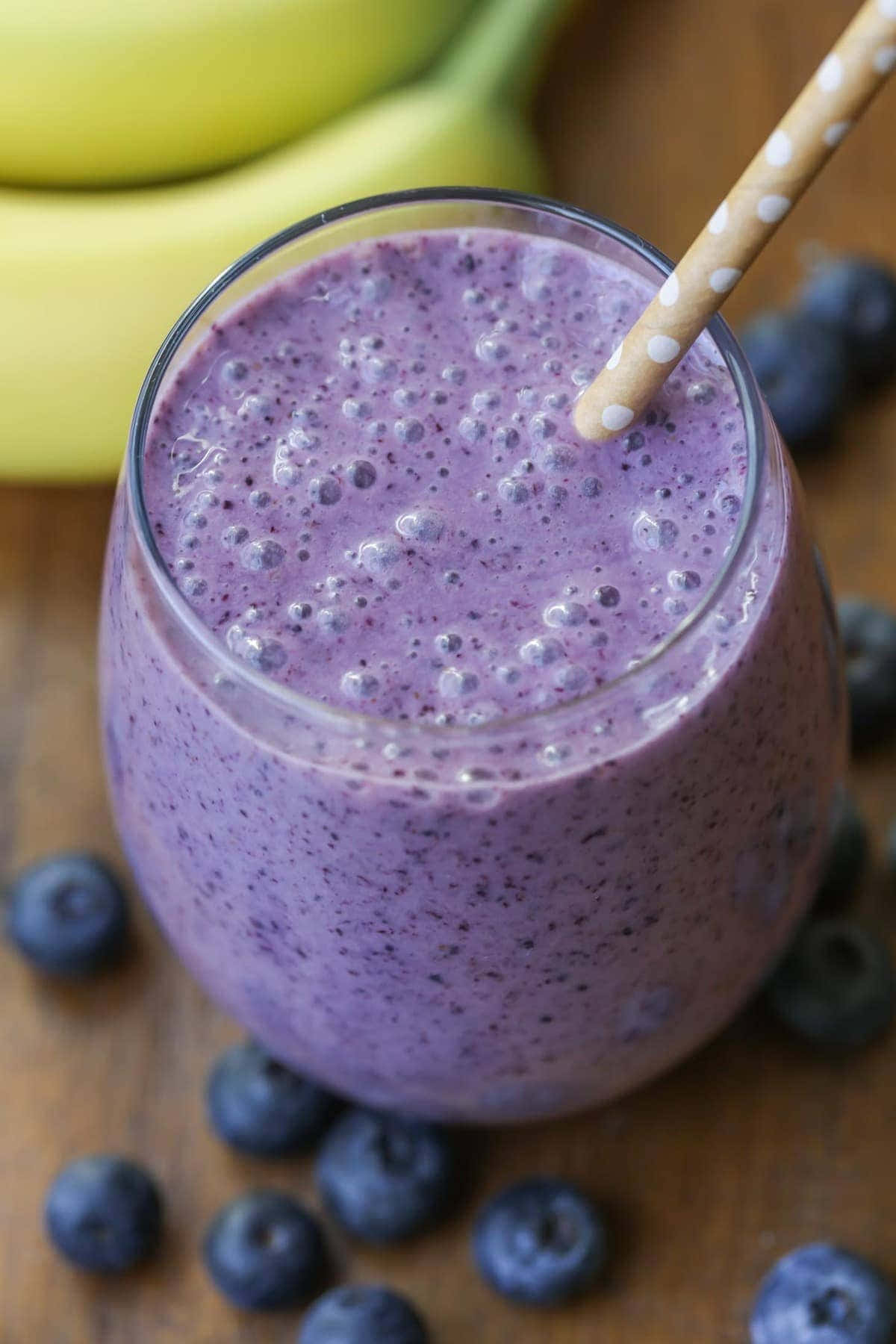 Blueberry Smoothie 桌布 壁紙 — 一杯充滿活力的紫藍色藍莓香蕉冰沙，放在玻璃杯中，插著波點吸管，周圍是新鮮藍莓和香蕉，置於木質表面上。