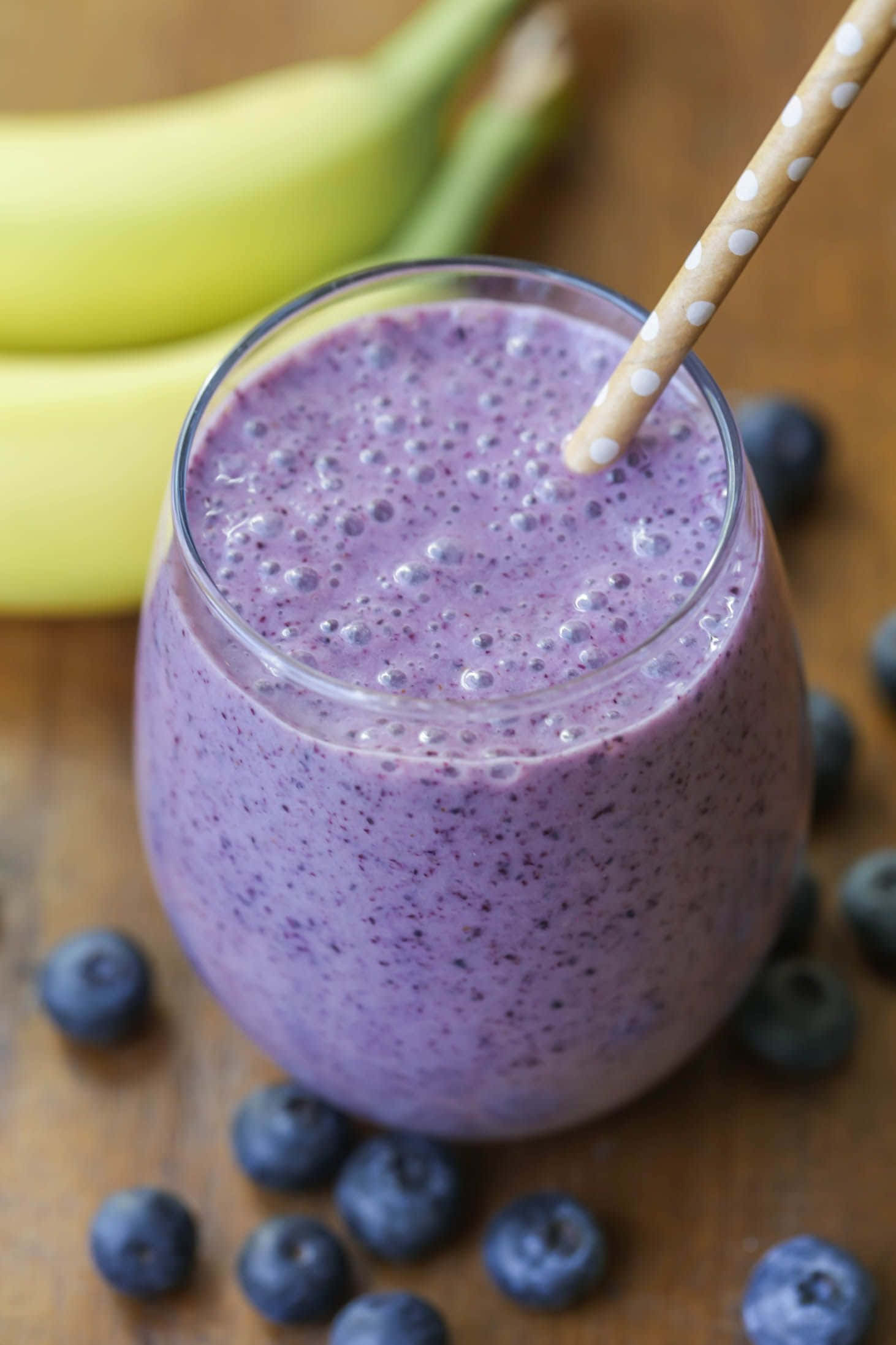 Blueberry Smoothie 桌布 壁紙 — 一杯充滿活力的紫藍莓香蕉冰沙，放在玻璃杯中，配上波點吸管，周圍是新鮮藍莓和香蕉，置於木質表面上。