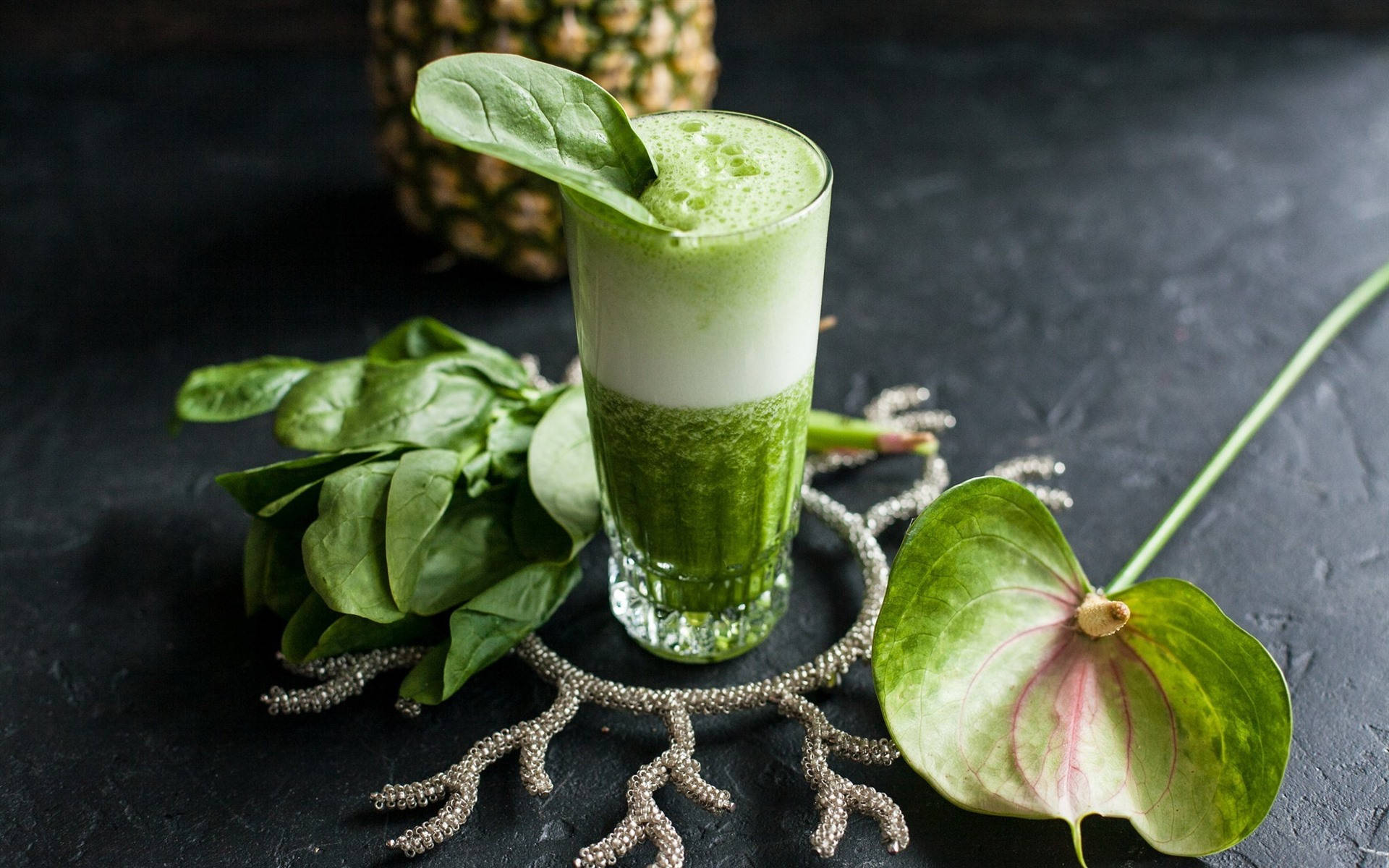 Smoothie 桌布 壁紙 — 一杯充滿活力的綠色冰沙，飾以菠菜葉，旁邊是新鮮菠菜、鳳梨和火鶴花，置於深色紋理表面上。