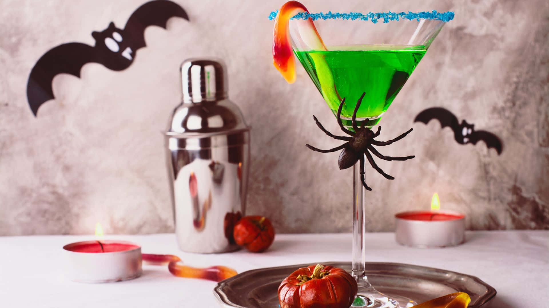 Spooky Cocktails 桌布 壁紙 — 一杯充滿活力的綠色萬聖節雞尾酒，盛載於馬丁尼杯中，飾有藍色邊糖果蜘蛛和橙色糖果裝飾。