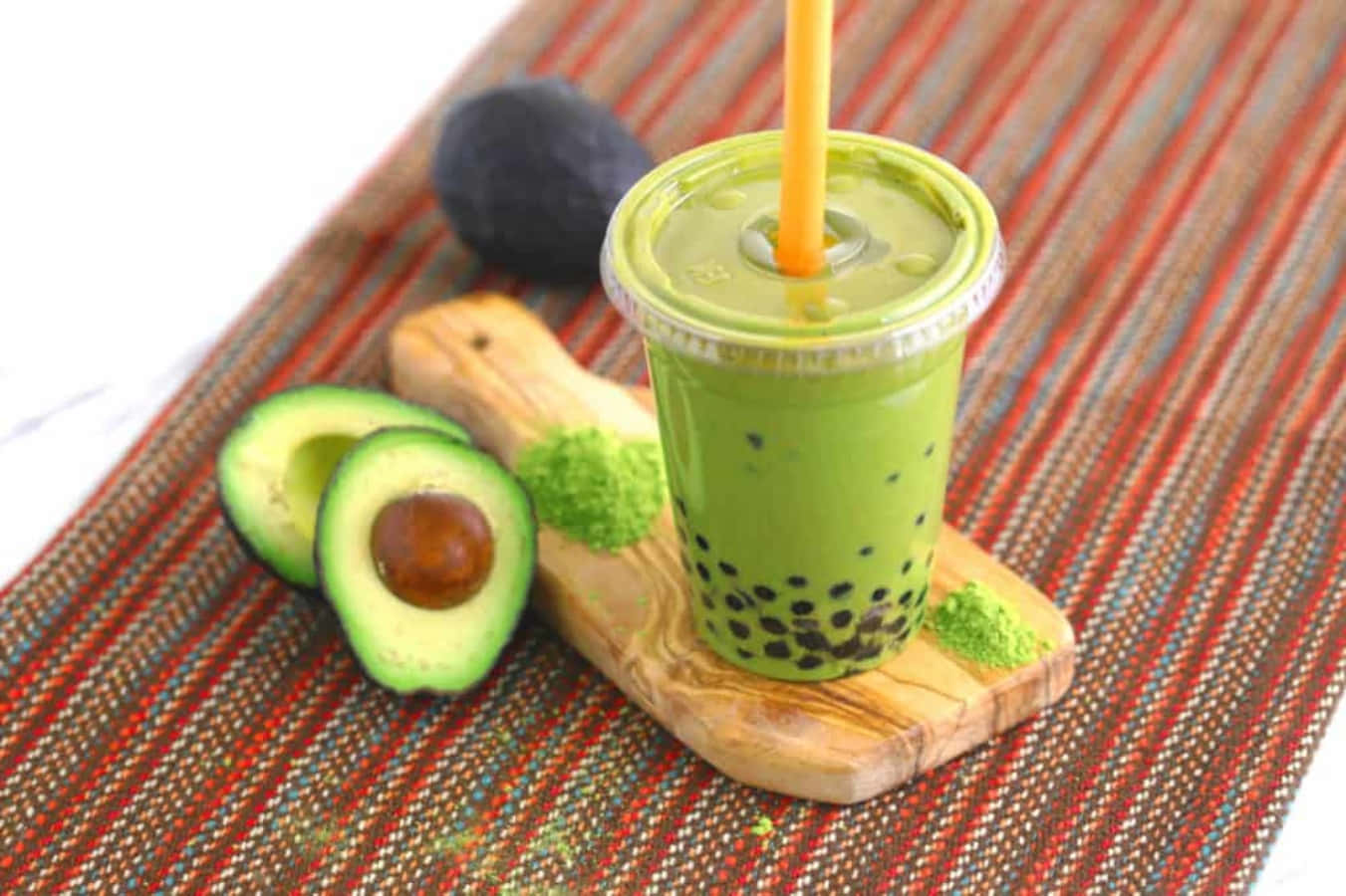 Boba Tea 桌布 壁紙 — 一杯充滿活力的綠色酪梨泡泡茶，放在透明杯中，搭配橘色吸管，置於木板上，旁邊有切片酪梨和粉末在條紋布上。