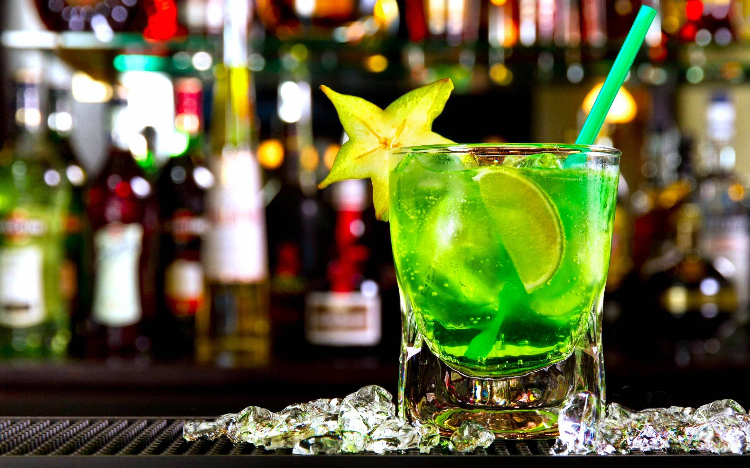 Drink 桌布 壁紙 — 一杯充滿活力的綠色雞尾酒，配上青檸和楊桃裝飾，放在碎冰上，背景是燈光昏暗的酒吧，後面有模糊的酒瓶。