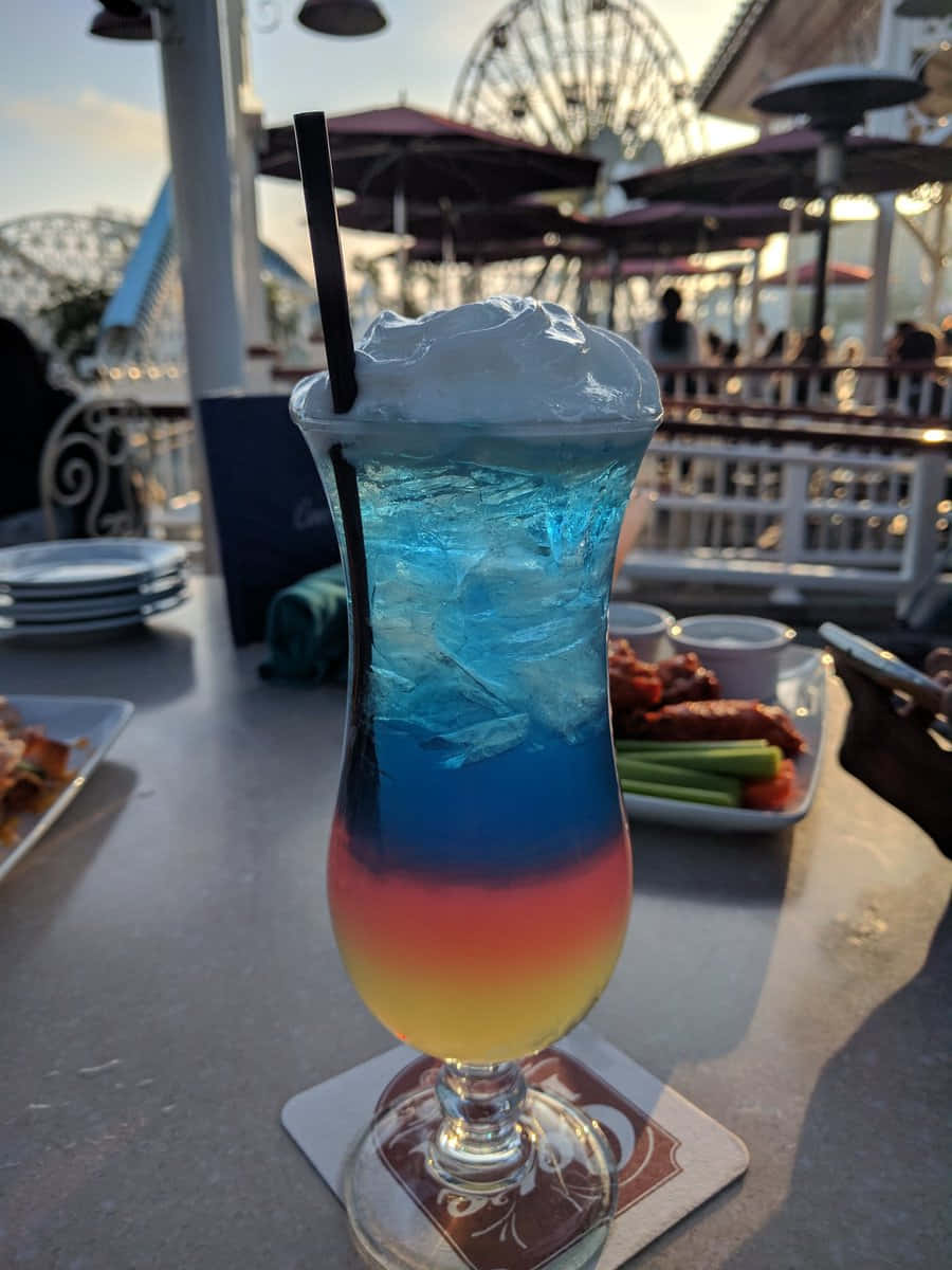Beautiful Drink 桌布 壁紙 — 一杯充滿活力的藍、紅、黃分層雞尾酒，配上鮮奶油和吸管，放在主題樂園戶外庭院的杯墊上，背景有摩天輪。