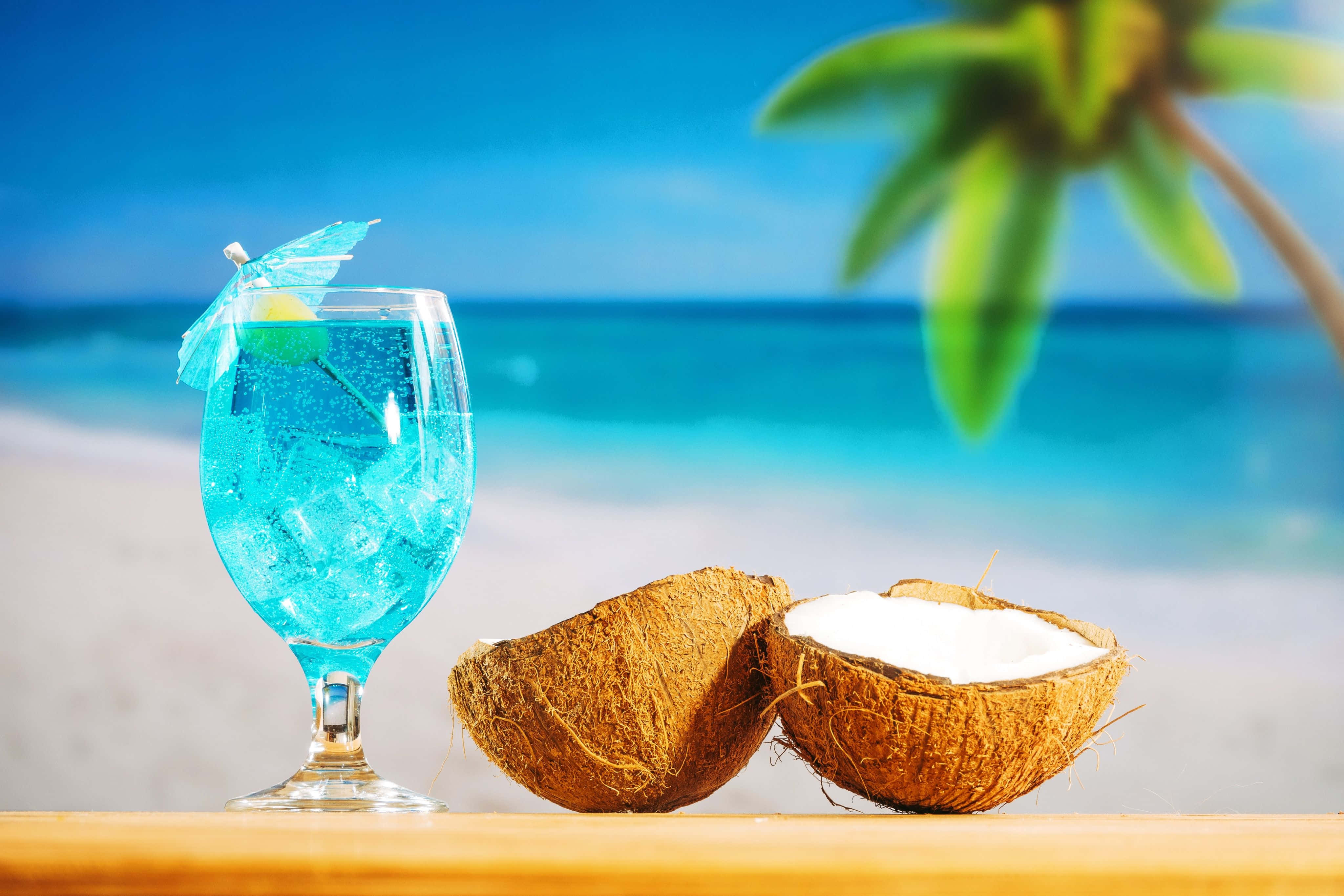 Coconut 桌布 壁紙 — 一杯充滿活力的藍色熱帶雞尾酒，配有冰塊和傘，放在木桌上，旁邊是一個裂開的椰子，背景是沙灘。